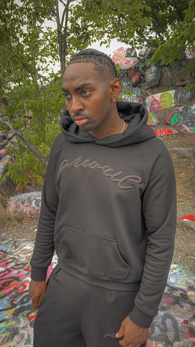 A 290-degree angled candid shot of a 6’1 light-skinned model with butterscotch-complexioned skin and reddish amber-brown dreads at the Quincy quarries, captured looking up at an elevated angle while wearing the full Onyx Set. The perspective highlights the 420 GSM heavyweight cotton-blend Onyx Hoodie, complimented by drop-shoulder saddled-raglan sleeve paneling, 1x1 rib-knit wrist cuffs and waistband, silk-threaded tonal "ONYX" embroidery across the chest and lower wrist, and an interior custom woven "V₂" n