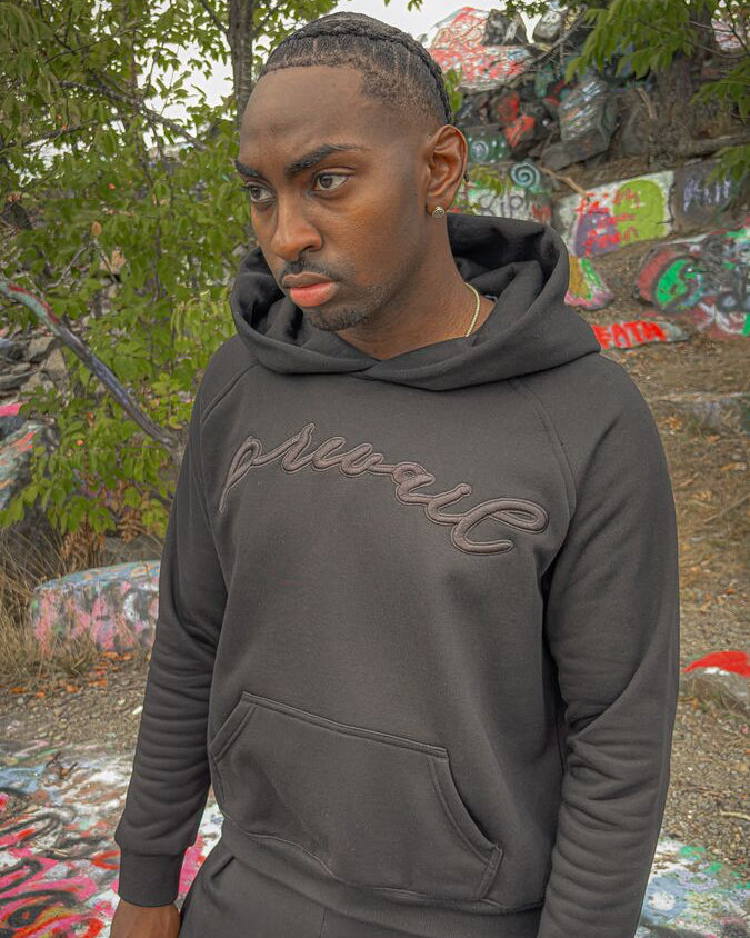 A 290-degree angled candid shot of a 6’1 light-skinned model with butterscotch-complexioned skin and reddish amber-brown dreads at the Quincy quarries, captured looking up at an elevated angle while wearing the full Onyx Set. The perspective highlights the 420 GSM heavyweight cotton-blend Onyx Hoodie, complimented by drop-shoulder saddled-raglan sleeve paneling, 1x1 rib-knit wrist cuffs and waistband, silk-threaded tonal "ONYX" embroidery across the chest and lower wrist, and an interior custom woven "V₂" 
