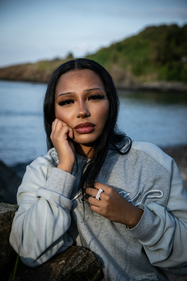 An on-body candid shot of a 5’7 female model looking at the lens front of the rocks on the beachside shoreline in Nahant, Massachusetts. Featuring the “emo” hoodie complimented by a saddled raglan-sleeve with drop shoulders, silk-threaded embroidery, French Terry paneling and a Heathered grey torso. Details include a distressed/frayed “ghost” kangaroo pocket, silk-thread torso embroidery, two-tone sleeve paneling, a saddled-raglan sleeve, and screen-printed silk woven tags.