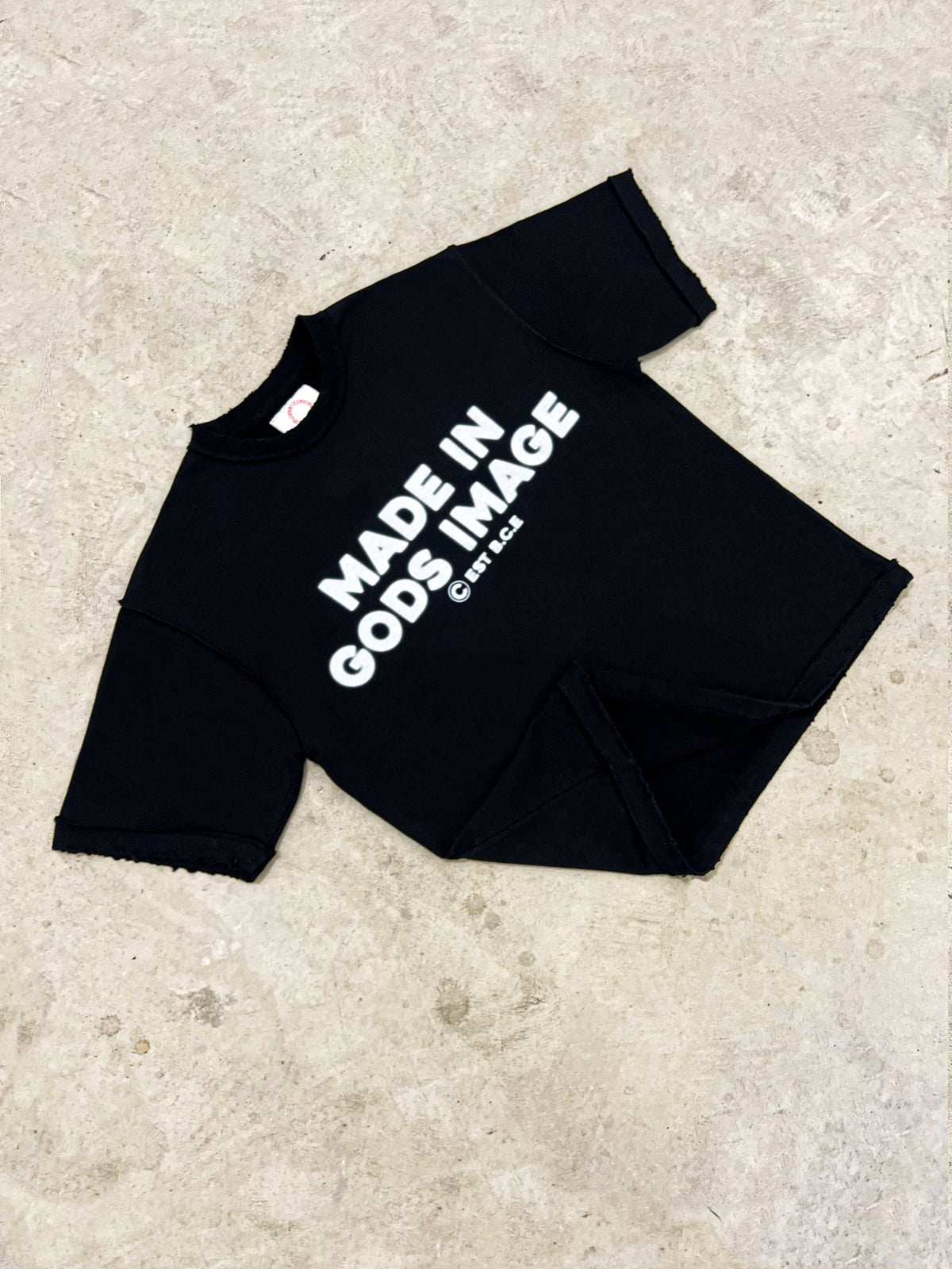 An angled flat lay of the MUST PREVAIL black 425 GSM heavyweight French Terry inside-out tee on a concrete surface. This shot highlights the 'MADE IN GODS IMAGE' white text, the oversized boxy silhouette, and the raw distressed hems.