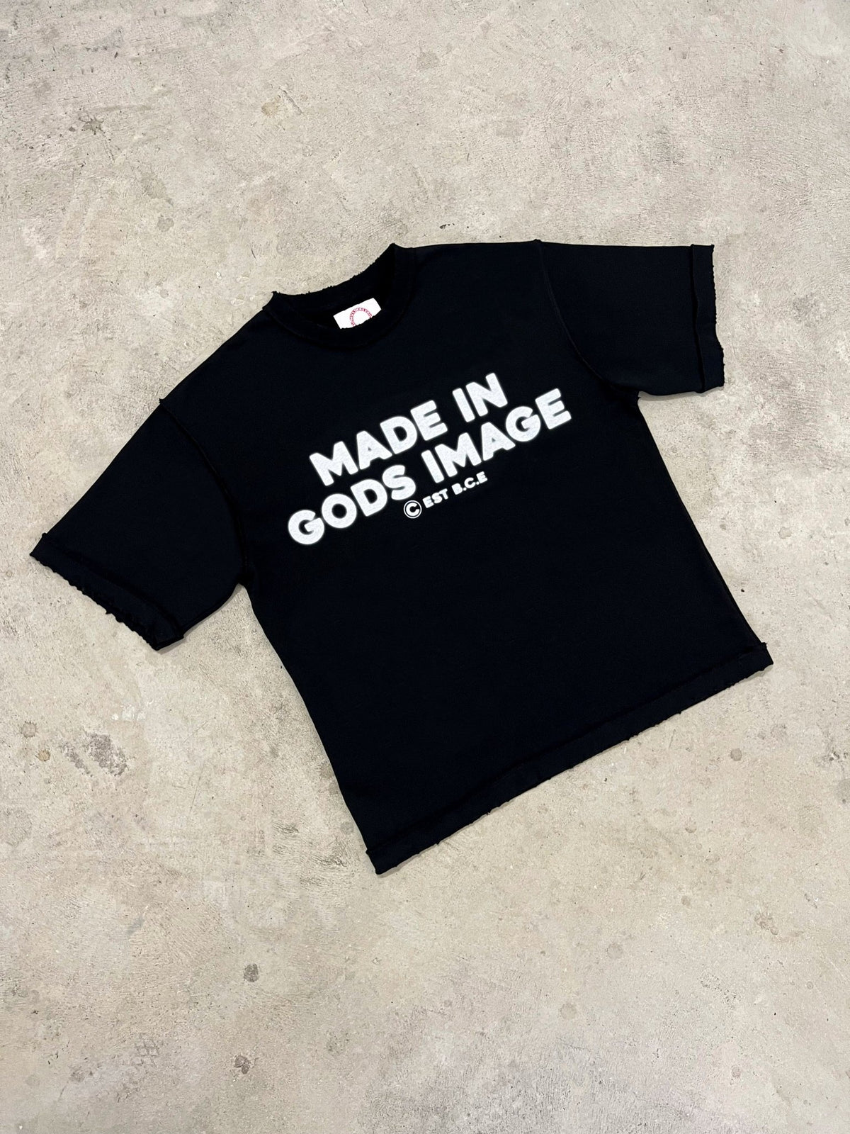 An angled flat lay of the MUST PREVAIL black 425 GSM heavyweight French Terry inside-out tee on a concrete background. This perspective highlights the 'MADE IN GODS IMAGE' white text, the oversized boxy silhouette, and the raw distressed hems. Showcases the premium fabric weight and technical construction of the garment.