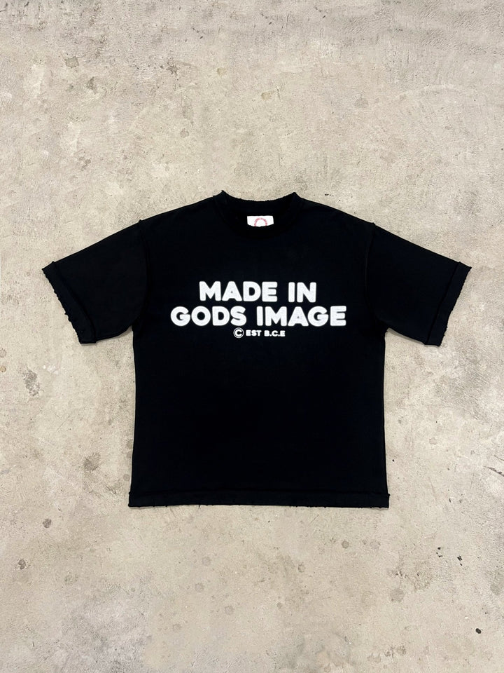 A flat lay of the MUST PREVAIL black 425 GSM heavyweight French Terry inside-out tee on a concrete surface. This shot highlights the 'MADE IN GODS IMAGE' white chest print, the oversized boxy fit, and the raw distressed hems. The industrial background showcases the structural texture and technical stitching of the garment.