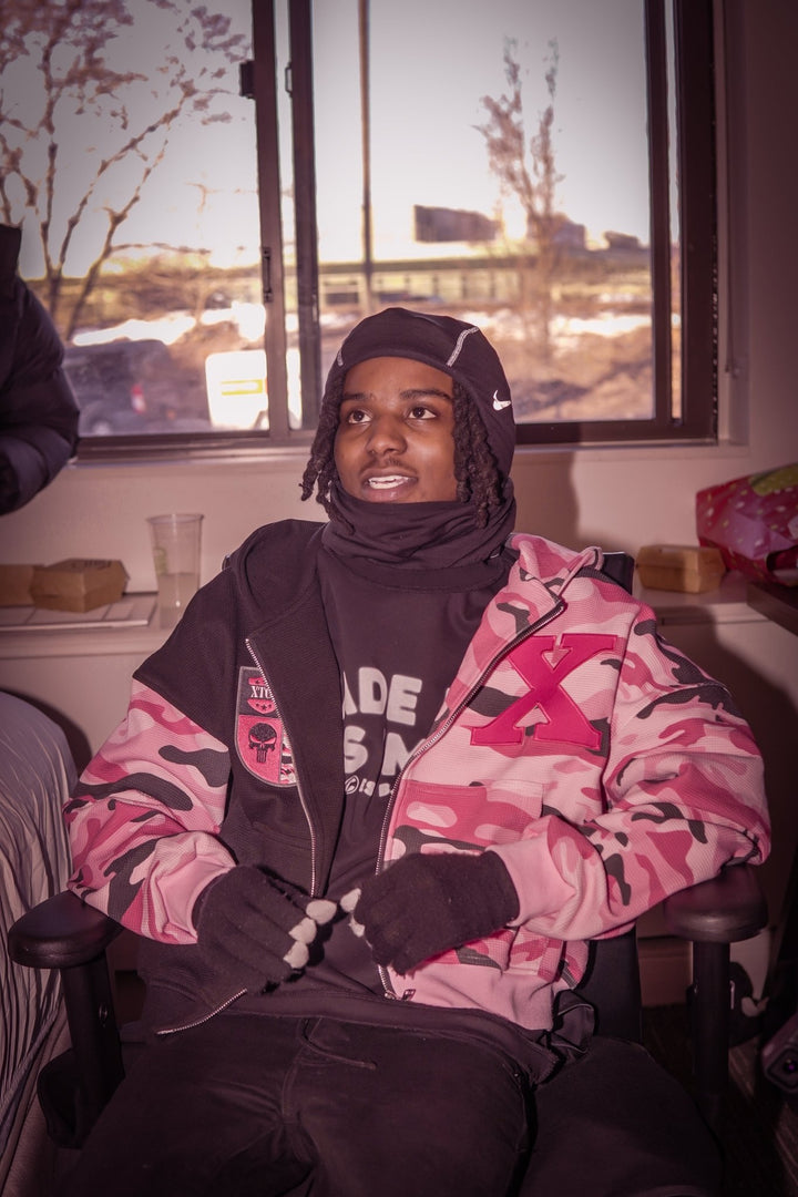 A candid lifestyle shot of a model wearing the MUST PREVAIL black 425 GSM heavyweight French Terry inside-out tee. Styled with a camo zip-up hoodie and a technical balaclava Nike ski mask, the image highlights how the oversized boxy fit maintains its structure while sitting. Features the 'MADE IN GODS IMAGE' chest print in a natural indoor setting.