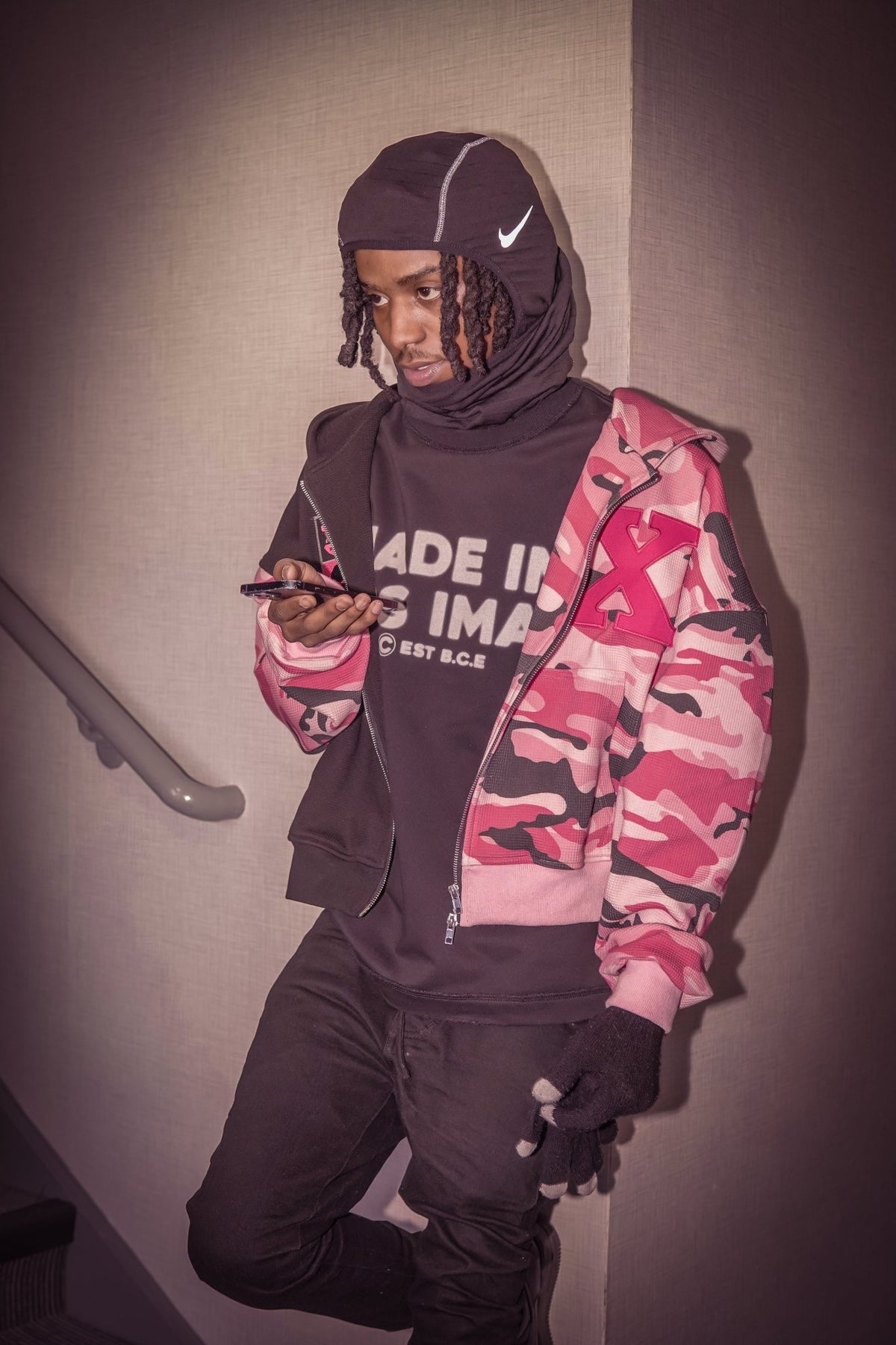 On-body lifestyle shot of the MUST PREVAIL black 425 GSM heavyweight French Terry inside-out tee. The model is layering the tee under a camo zip-up hoodie with a technical balaclava Nike ski mask, showcasing the boxy oversized fit and structural 'MADE IN GODS IMAGE' chest print in a high-contrast indoor setting.