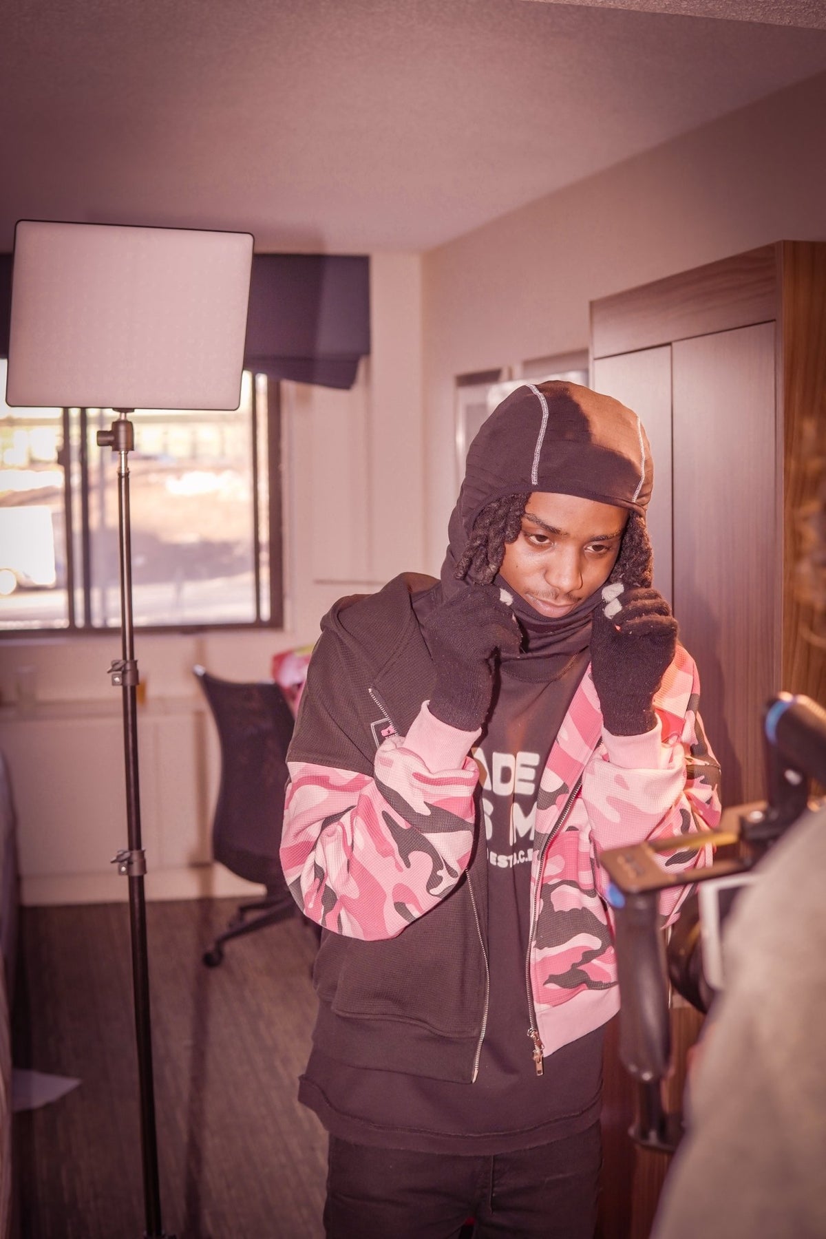 A behind-the-scenes look at a MUST PREVAIL studio shoot featuring the black 425 GSM heavyweight French Terry inside-out tee. The model is layering the tee under a camo zip-up, styled with a technical balaclava Nike ski mask. Lighting equipment and camera gear are visible, highlighting the professional production and boxy fit of the garment.