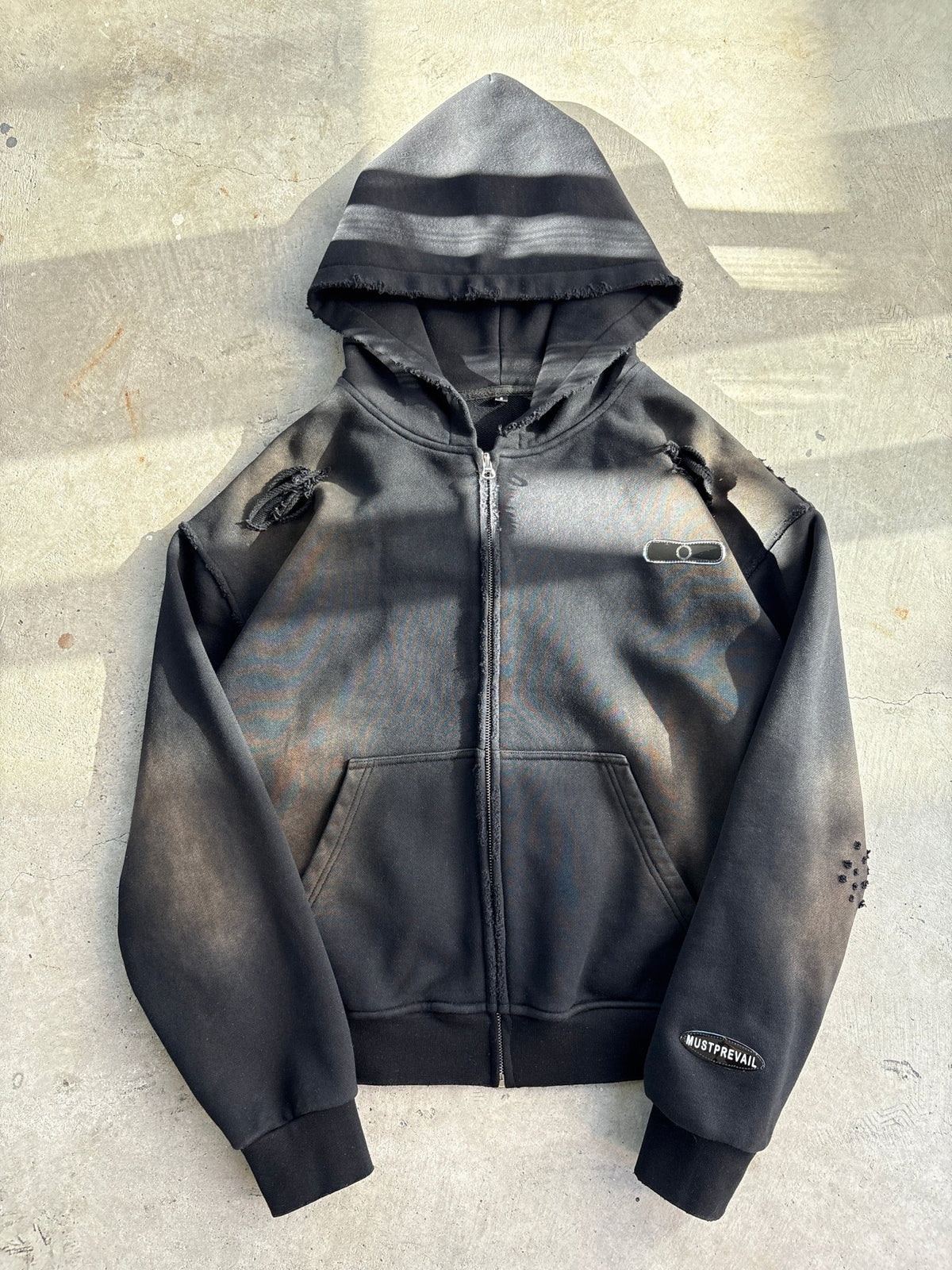 A flat lay of a black 500 GSM heavyweight French Terry full-zip hoodie on a concrete surface. The garment features a cropped-drop shoulder fit, oversized deep-V hood, and YKK zipper. Details include a two-tone sand-blast vintage aesthetic with micro-distressing, sewn-on adhesive labeling, and screen-printed silk woven tags