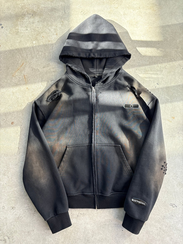 A flat lay of a black 500 GSM heavyweight French Terry full-zip hoodie on a concrete surface. The garment features a cropped-drop shoulder fit, oversized deep-V hood, and YKK zipper. Details include a two-tone sand-blast vintage aesthetic with micro-distressing, sewn-on adhesive labeling, and screen-printed silk woven tags