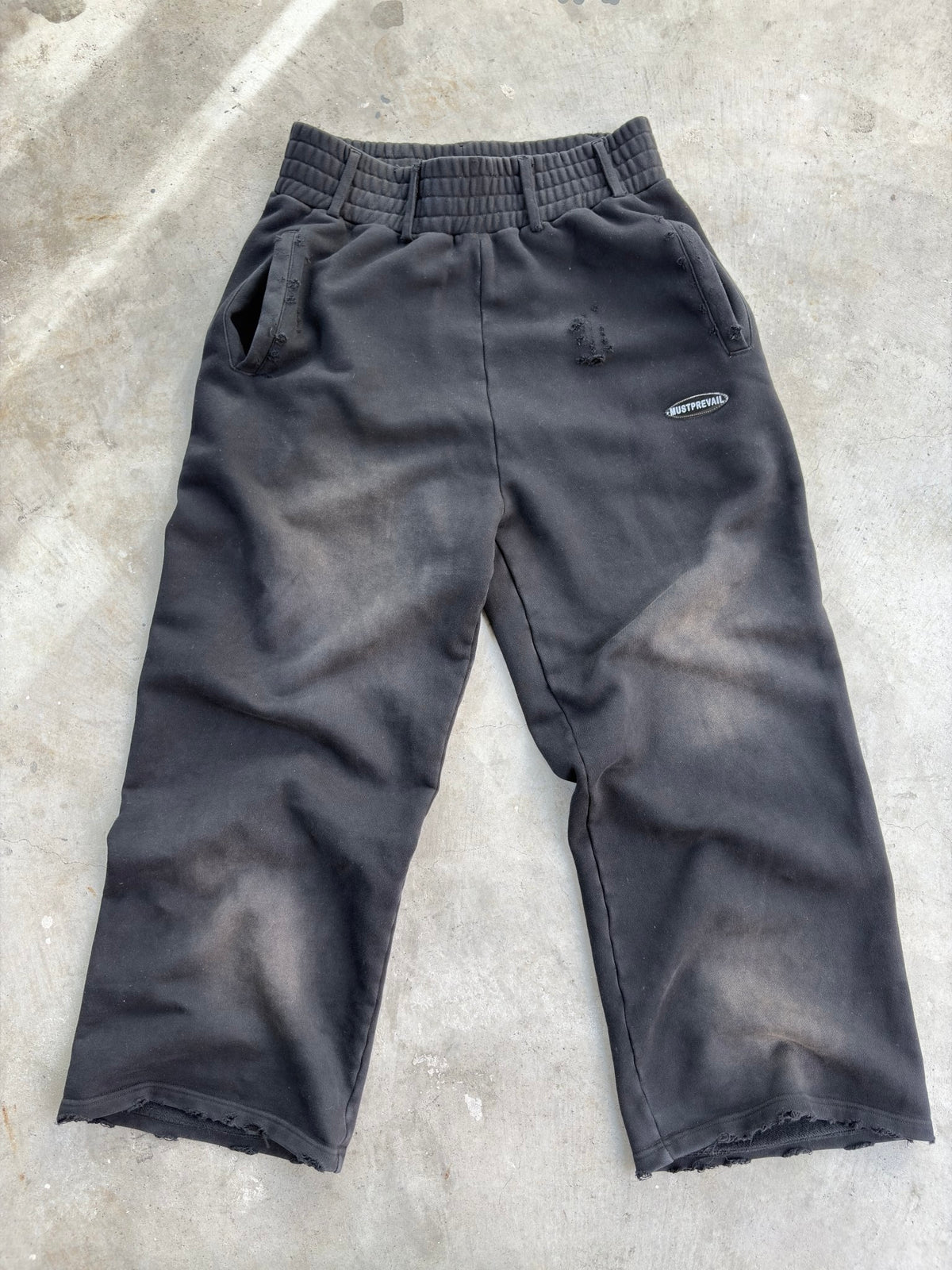 A flat lay of the black 500 GSM French Terry baggy sweatpants on a concrete surface. Showcases the unique triple waistband with belt loops, wide-leg fit, and two-tone sand-blast aesthetic. Features micro-distressing, double-front and back pockets, and sewn-on adhesive labeling on a heavy-weight Y2K-inspired silhouette.