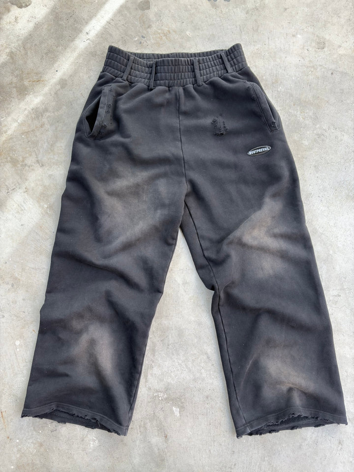 A flat lay of the black 500 GSM French Terry baggy sweatpants on a concrete surface. Showcases the unique triple waistband with belt loops, wide-leg fit, and two-tone sand-blast aesthetic. Features micro-distressing, double-front and back pockets, and sewn-on adhesive labeling on a heavy-weight Y2K-inspired silhouette.