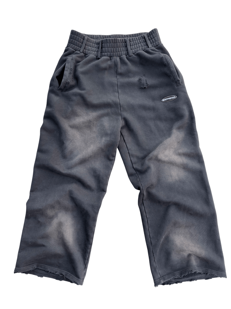 A transparent flat lay of the MUST PREVAIL black 500 GSM French Terry baggy sweatpants. Features a unique triple waistband with belt loops, a wide-leg fit, and 2-tone sand-blast distressing. Detailed with double-front and back pockets, sewn-on adhesive labeling, and screen-printed silk woven tags on a heavyweight Y2K-inspired silhouette.