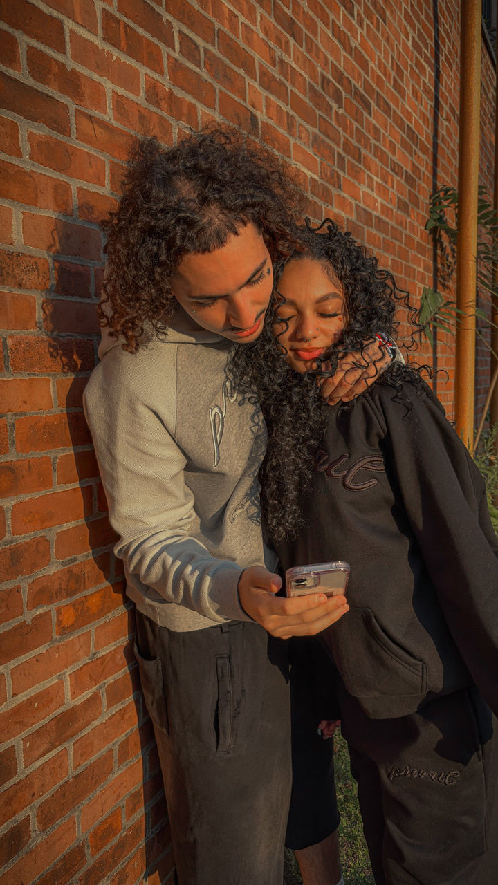 A candid medium shot of the same couple standing together in front of a red brick wall. The 5'6" female model in the black Onyx set holds her phone out as both she and the 6'1" male model in the Emo Hoodie look down at the screen, smiling while reviewing the photo they just took. The image captures the relaxed lifestyle mood alongside the distinct two-tone paneling of the Emo Hoodie and the clean structural drape of the Onyx set.