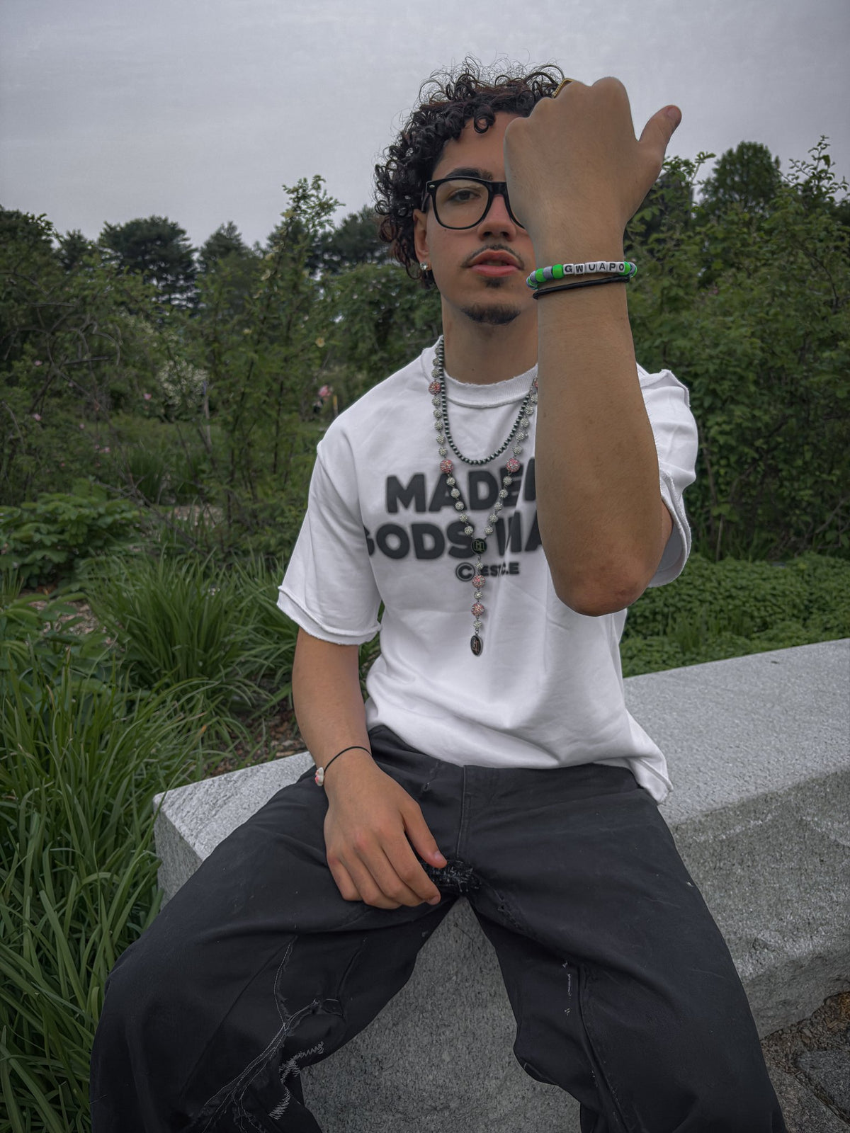 A relaxed candid photograph of the 6'0" light-skinned male model with curly brown hair, sitting casually on a large raw concrete barrier block in a lush green park. He wears the white colorway of the Inside Out Tee (425 GSM heavy-weight French Terry Cotton, oversized boxy fit), detailed with its characteristic reverse-seam construction and DTG print. He is styled with grayish-green utilitarian cargo jeans and cream-colored skate shoes, looking comfortably at the camera with a subtle expression. 