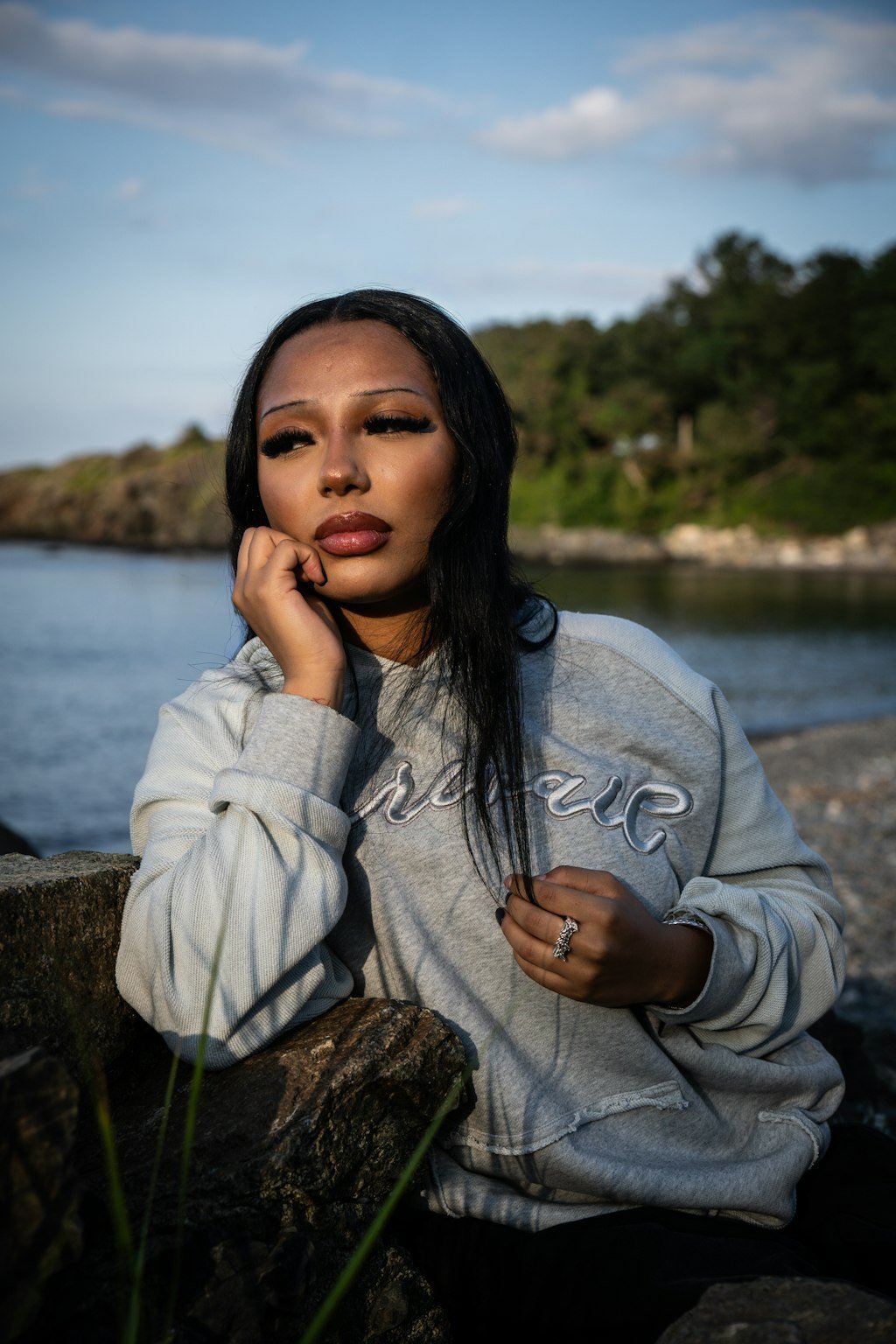 An on-body candid shot of a 5’7 female model sitting in front of the coastal rocks along the beachside shoreline in Nahant, Massachusetts, wearing the Emo Hoodie. The seated angle highlights the 420 GSM heavyweight French Terry cotton construction featuring a heathered grey torso, two-tone sleeve paneling, and saddled-raglan drop-shoulder construction. Detailed elements showcase the distressed and frayed deconstructed "ghost" kangaroo pocket, silk-threaded tonal torso embroidery, 2x2 rib-knit cuffs and wais