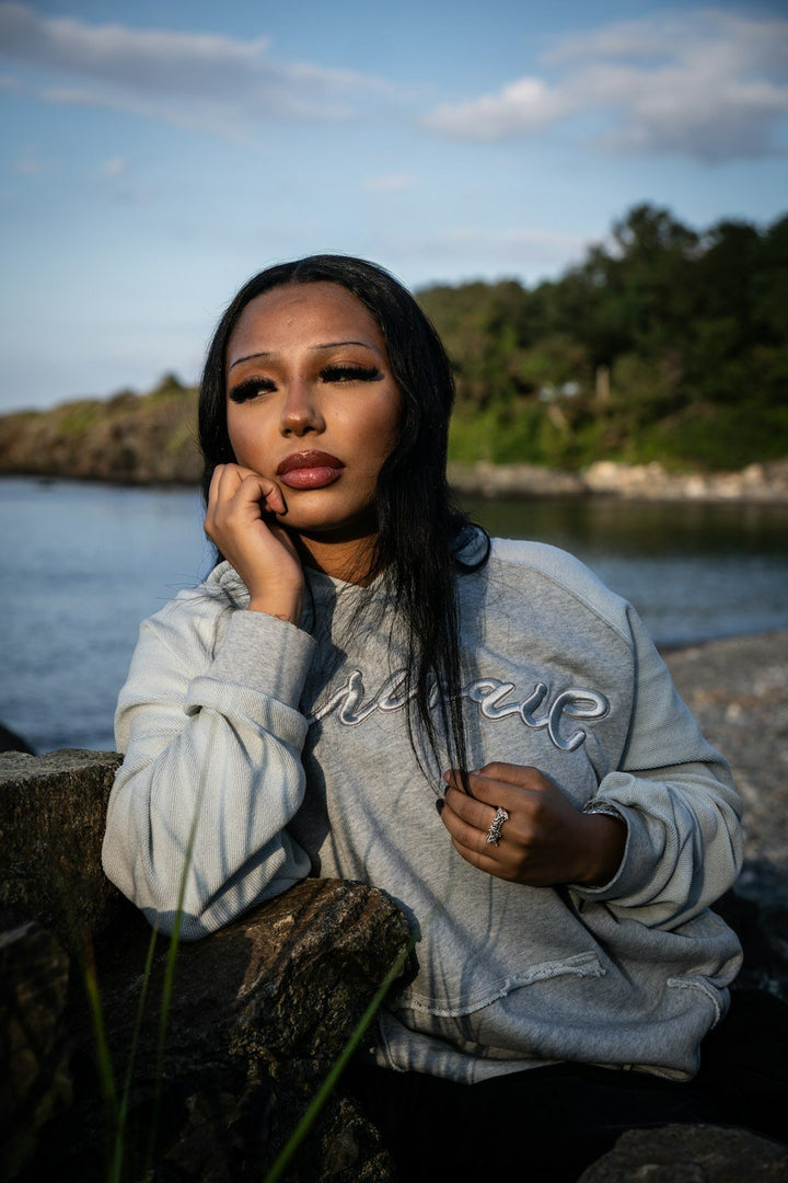 An on-body candid shot of a 5’7 female model sitting in front of the coastal rocks along the beachside shoreline in Nahant, Massachusetts, wearing the Emo Hoodie. The seated angle highlights the 420 GSM heavyweight French Terry cotton construction featuring a heathered grey torso, two-tone sleeve paneling, and saddled-raglan drop-shoulder construction. Detailed elements showcase the distressed and frayed deconstructed "ghost" kangaroo pocket, silk-threaded tonal torso embroidery, 2x2 rib-knit cuffs and wais
