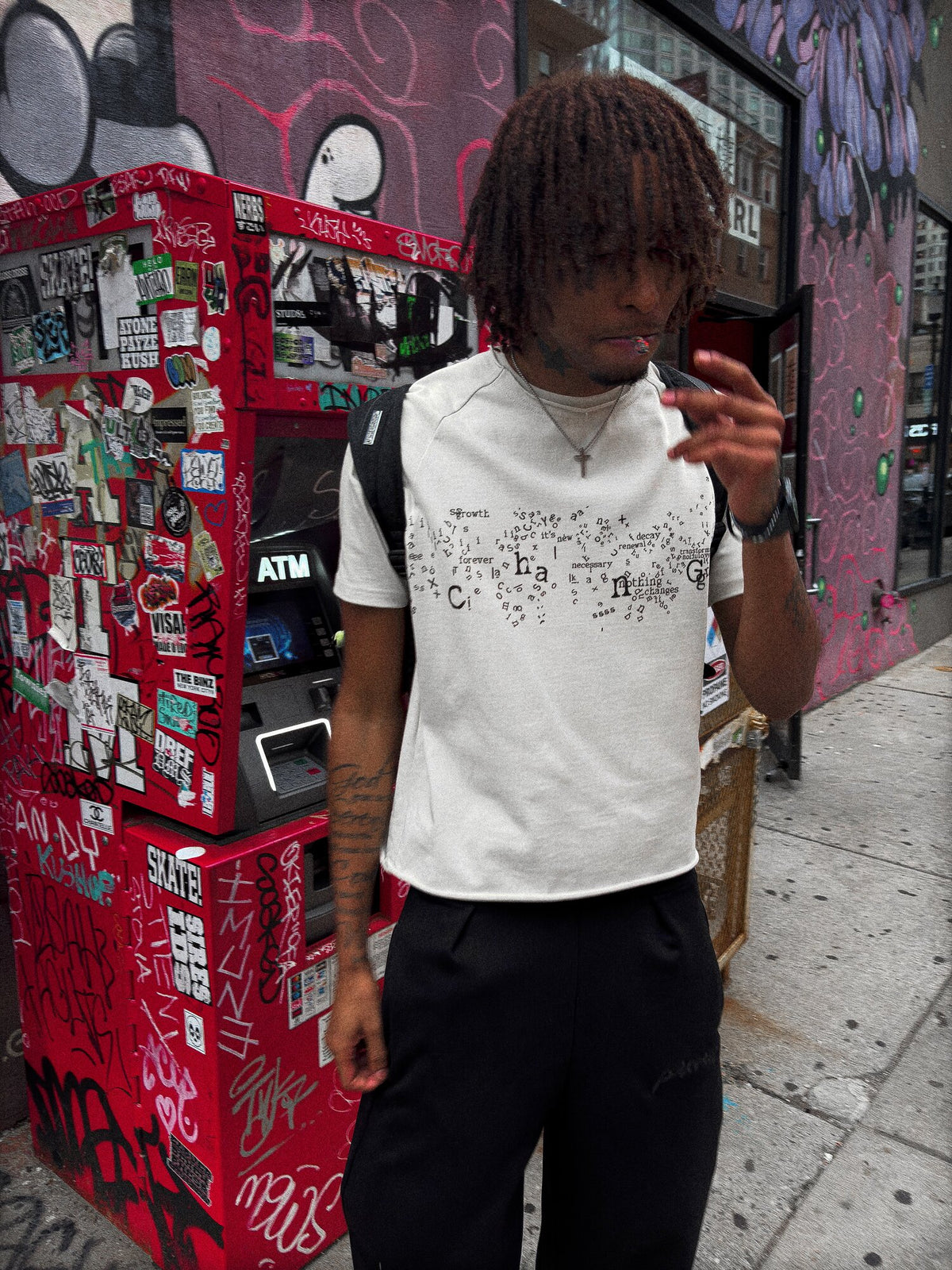 An on-body shot of a 6’1 light-skinned model with butterscotch-complexioned skin and reddish amber-brown dreads, captured candidly while smoking in front of the Chinatown gate ATM in Boston, wearing the "poem" tee paired with the Onyx Sweats. The top showcases the 325 GSM French Terry fleece-cotton tee, complimented by saddled-raglan sleeve paneling with a slim drop-shoulder fit, a direct-to-garment torso print, a stacked raw-edge ribbed neckline, and subtle hand-cut Dremel micro-distressing throughout. The