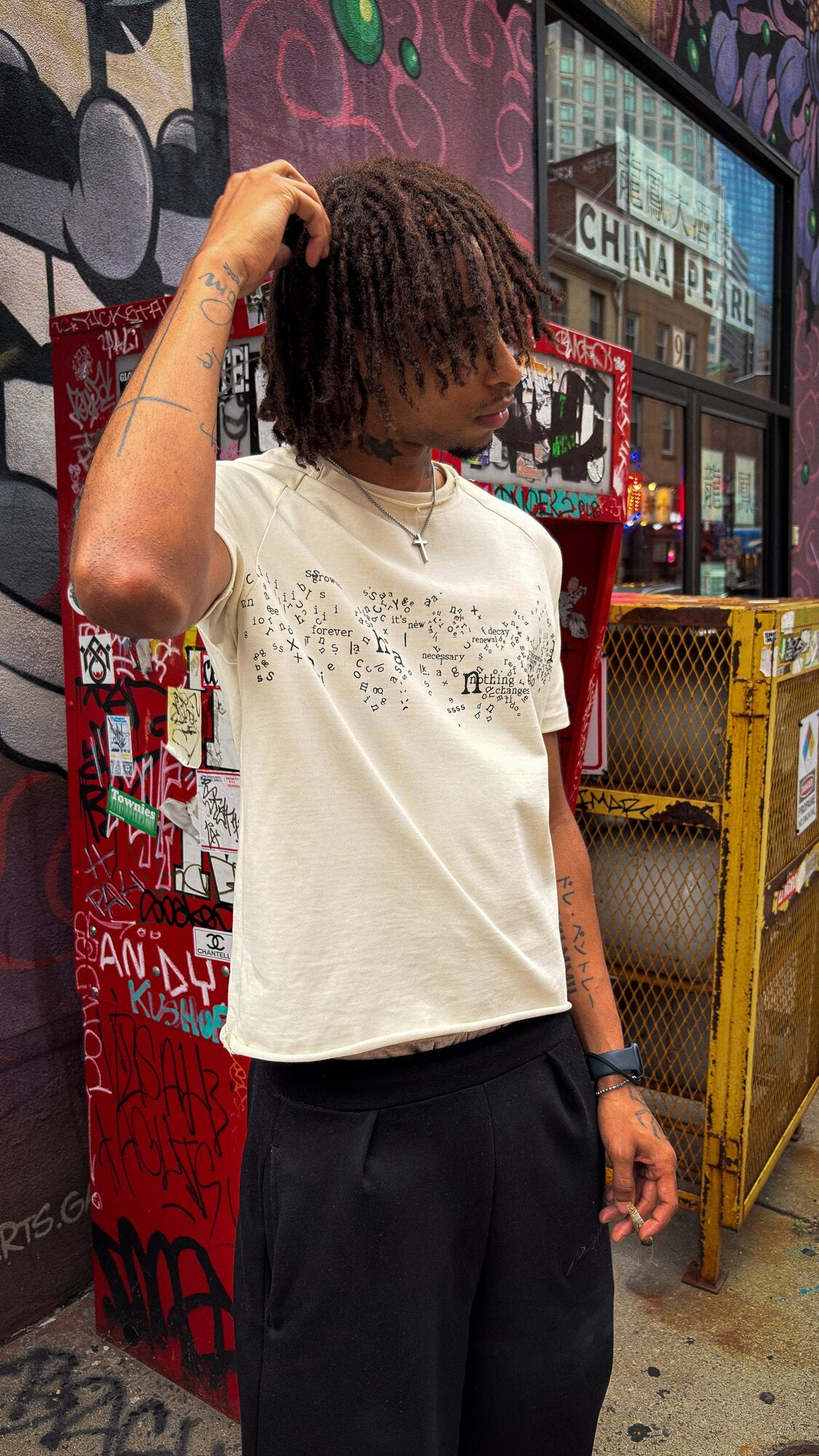 An on-body shot of a 6’1 light-skinned model with butterscotch-complexioned skin and reddish amber-brown dreads, standing in front of the iconic Chinatown gate ATM in Boston while wearing the "poem" tee paired with the Onyx Sweats. The top showcases the 325 GSM French Terry fleece-cotton tee, complimented by saddled-raglan sleeve paneling with a slim drop-shoulder fit, a direct-to-garment torso print, a stacked raw-edge ribbed neckline, and subtle hand-cut Dremel micro-distressing throughout. The bottom hig