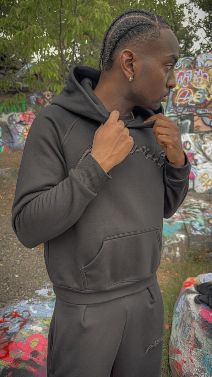 An on-body candid shot of a 6’1 male model standing in front of the historic stone Quincy barracks beside large graffiti-covered rocks, wearing a size medium Onyx Set. The top features the 420 GSM heavyweight cotton-blend Onyx Hoodie, complimented by drop-shoulder saddled-raglan sleeve paneling, 1x1 rib-knit wrist cuffs and waistband, silk-threaded tonal "ONYX" embroidery across the chest and lower wrist, a seamless deconstructed "ghost" front kangaroo pocket, and an interior custom woven "V₂" neck tag. The
