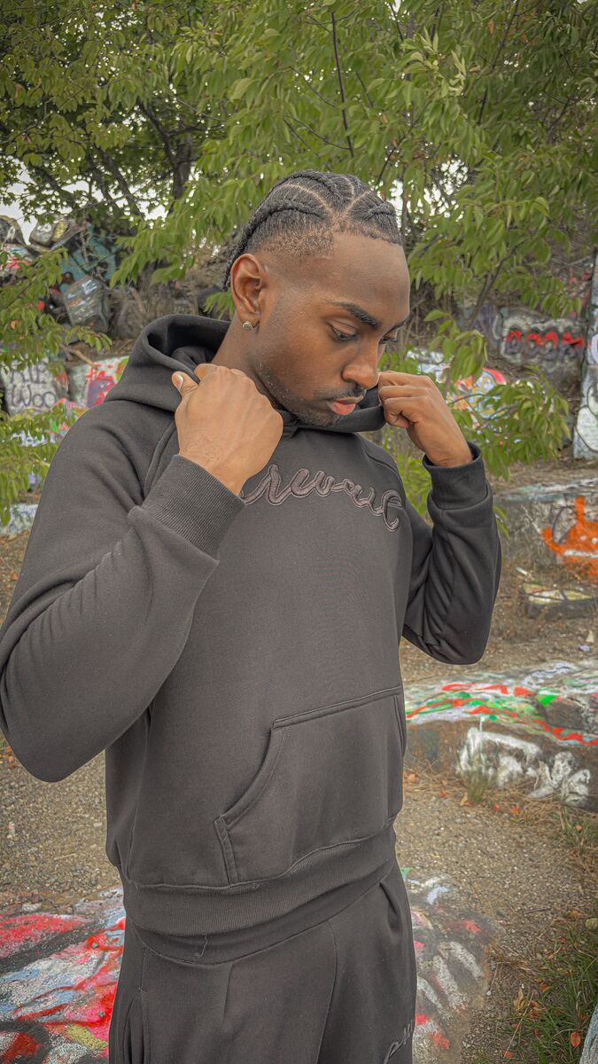 An on-body candid shot of a 6’1 male model standing in front of the historic stone Quincy barracks beside large graffiti-covered rocks, wearing a size medium Onyx Set with his head tilted down facing forward. The top features the 420 GSM heavyweight cotton-blend Onyx Hoodie, complimented by drop-shoulder saddled-raglan sleeve paneling, 1x1 rib-knit wrist cuffs and waistband, silk-threaded tonal "ONYX" embroidery across the chest and lower wrist, a seamless deconstructed "ghost" front kangaroo pocket, and an