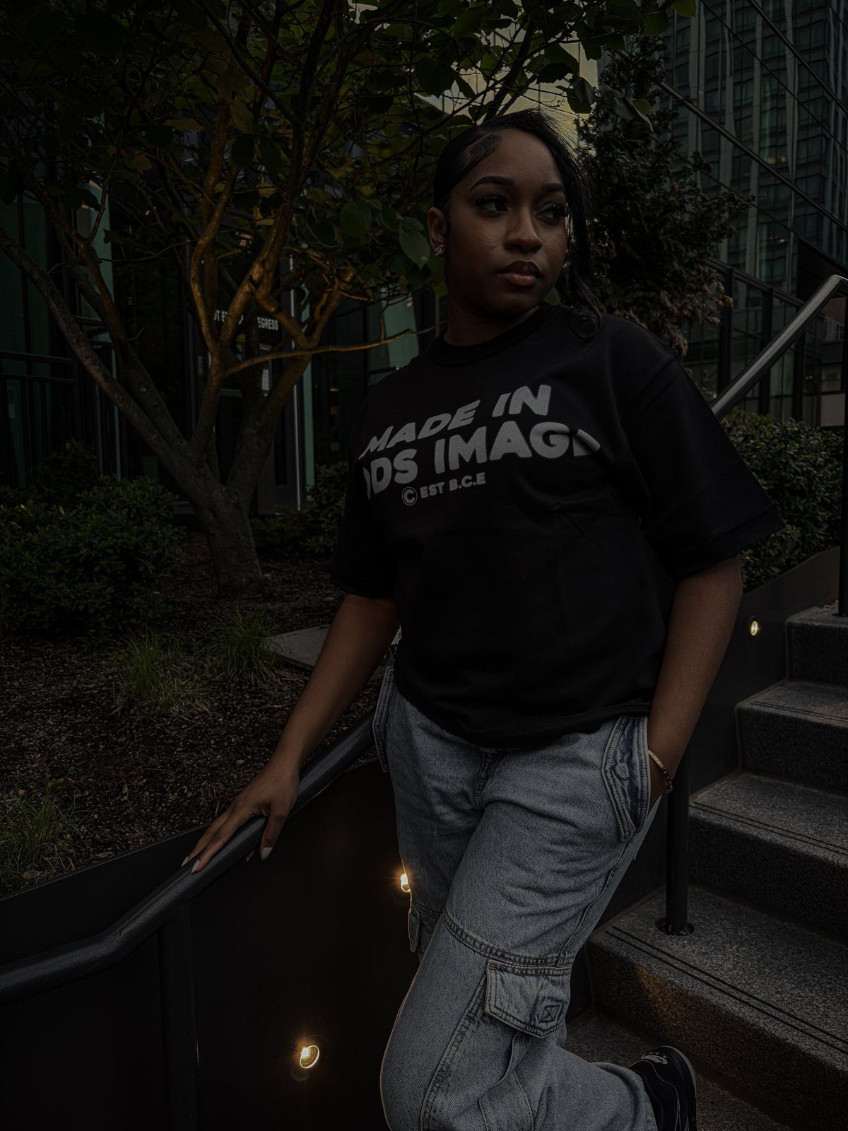 A candid outdoor photograph, captured in soft daylight on the iron railing of brownstone steps in front of a leafy green tree. The 5'9" female model wears the black Inside Out Tee (425 GSM heavy-weight French Terry Cotton, oversized boxy fit), showcasing its reverse-seam construction and direct-to-garment (DTG) print on the chest. She has dark, wavy hair and is paired with classic medium-wash straight-leg jeans and black canvas sneakers, leaning casually on the railing with her legs crossed.