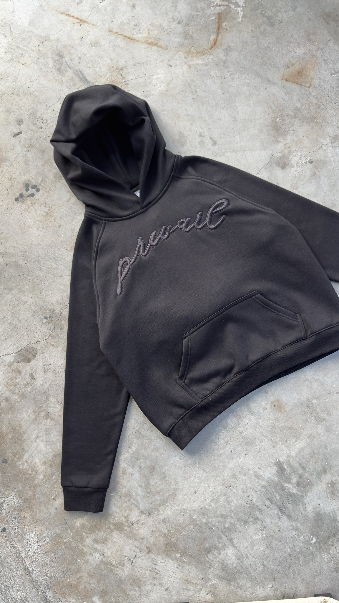 A top-down diagonal flat lay shot taken from an 8-foot overhead angle on a concrete garage floor, capturing the Onyx Hoodie positioned at a tilted angle pointing toward the top-left corner with sleeves spread wide to showcase overall garment layout profiling and proportions. The detailed perspective highlights the 420 GSM heavyweight cotton-blend construction, featuring saddled-raglan drop-shoulder sleeve paneling, 1x1 rib-knit wrist cuffs and waistband, silk-threaded tonal torso and wrist embroidery