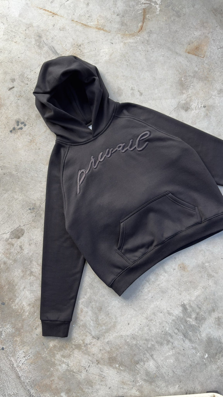 A top-down diagonal flat lay shot taken from an 8-foot overhead angle on a concrete garage floor, capturing the Onyx Hoodie positioned at a tilted angle pointing toward the top-left corner with sleeves spread wide to showcase overall garment layout profiling and proportions. The detailed perspective highlights the 420 GSM heavyweight cotton-blend construction, featuring saddled-raglan drop-shoulder sleeve paneling, 1x1 rib-knit wrist cuffs and waistband, silk-threaded tonal torso and wrist embroidery
