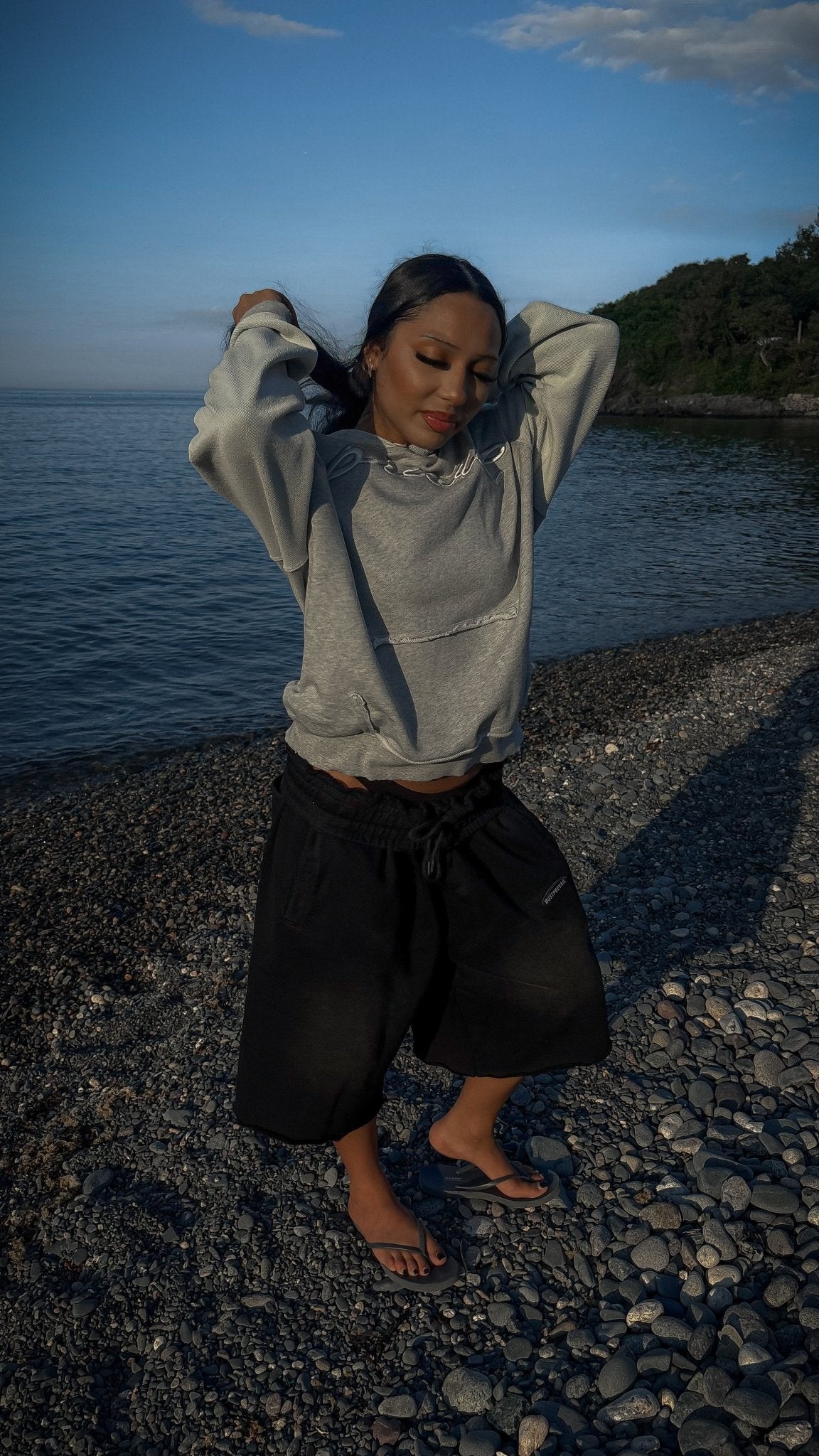 An editorial on-body shot of a 5’7 female model standing on coastal rocks in front of the ocean water along the beachside shoreline in Nahant, Massachusetts, looking down while holding her hair. The atmospheric framing highlights the 420 GSM heavyweight French Terry cotton construction of the Emo Hoodie, featuring a heathered grey torso, two-tone sleeve paneling, and saddled-raglan drop-shoulder construction. Key details highlight the distressed and frayed deconstructed "ghost" kangaroo pocket, silk-threade