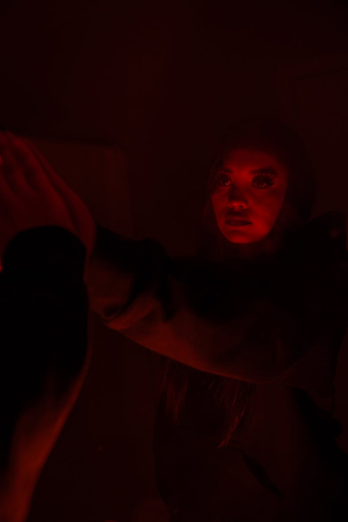 An editorial shot of a 5’7 female model in a dimly lit bathroom featuring ambient red lighting against a dark background, highlighting the contrast and silhouette of the black Onyx Hoodie. The moody perspective showcases the 420 GSM heavyweight cotton-blend construction, complimented by drop-shoulder saddled-raglan sleeve paneling and 1x1 rib-knit cuffs and waistband. Details include tonal silk-threaded embroidery across the chest and lower wrist, alongside an interior custom woven "V₂" neck tag.