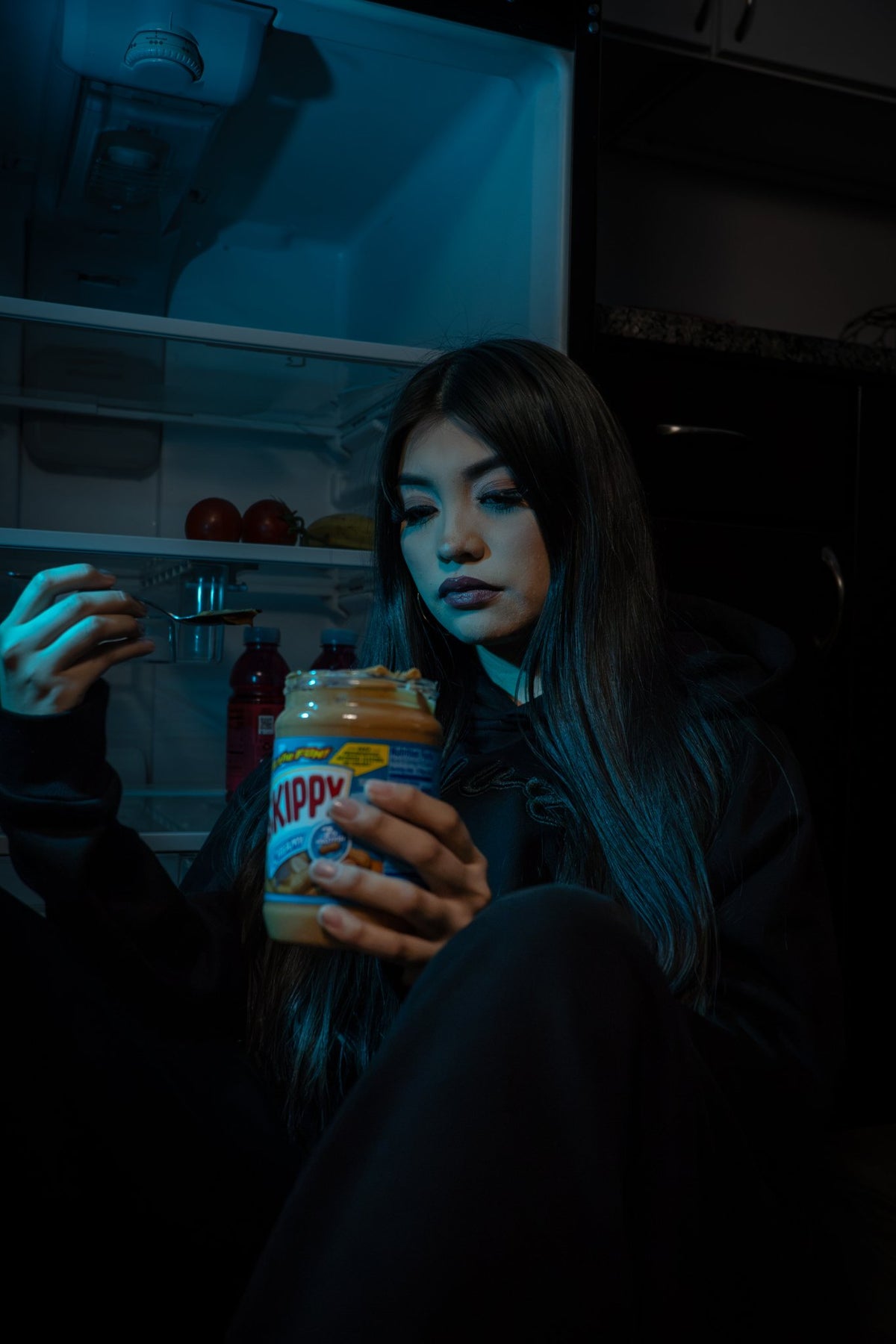 An editorial shot of a 5’7 female model sitting down in front of a refrigerator cast in ambient amber and blue light, wearing the complete Onyx Set. The top highlights the 420 GSM heavyweight cotton-blend Onyx Hoodie, complimented by drop-shoulder saddled-raglan sleeve paneling, 1x1 rib-knit wrist cuffs and waistband, silk-threaded tonal "ONYX" embroidery across the chest and lower wrist, and an interior custom woven "V₂" neck tag. The bottom features matching 420 GSM true-to-size baggy wide-leg Onyx Sweats