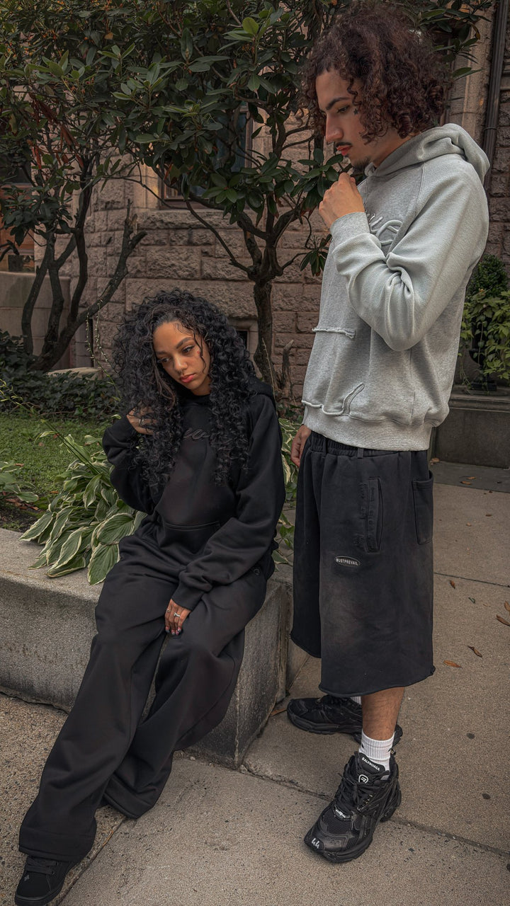 An editorial full-length outdoor shot on the Newbury Street sidewalk. On the left, a 5'6" female model poses wearing the complete black Onyx Hoodie and Onyx Sweats set (420 GSM heavyweight cotton blend with tonal silk embroidery). Beside her, a 6'1" light-skinned male model with curly hair wears the Emo Hoodie (420 GSM French Terry with heathered grey torso and ghost pocket details) paired with unreleased Y2K 3/4 length shorts. The composition highlights the structured streetwear silhouettes.