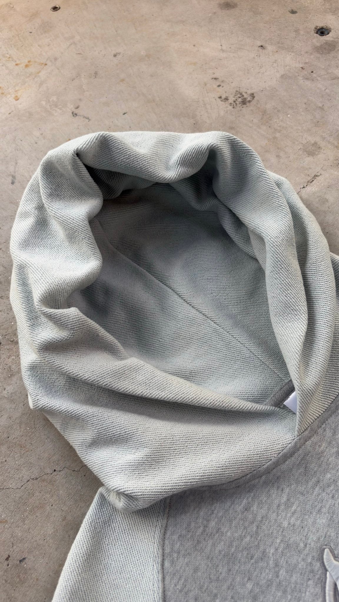 A top-down flat lay shot of the Emo Hoodie spread diagonally across a raw concrete garage floor with the hood pointing toward the left corner. The angled perspective showcases the structural silhouette and high-grade 420 GSM heavyweight French Terry cotton loops, detailing the heavy fabric density, two-tone sleeve paneling, saddled-raglan drop-shoulder construction, and the overall architectural geometry of the garment.