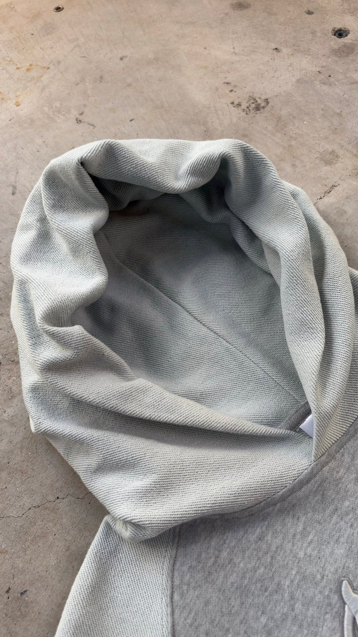 A top-down flat lay shot of the Emo Hoodie spread diagonally across a raw concrete garage floor with the hood pointing toward the left corner. The angled perspective showcases the structural silhouette and high-grade 420 GSM heavyweight French Terry cotton loops, detailing the heavy fabric density, two-tone sleeve paneling, saddled-raglan drop-shoulder construction, and the overall architectural geometry of the garment.