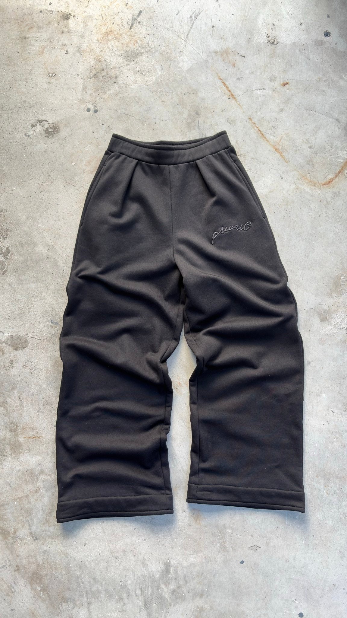 A zoomed-out overhead flat lay shot of the black Onyx Sweats laid out on a concrete garage floor, showcasing the full silhouette and wide-leg proportions at a 3/4 angle. The high-angle perspective details the 420 GSM heavyweight cotton-blend construction, non-roll tubular waistband finishing, tonal silk-threaded upper thigh embroidery, and true-to-size baggy fit geometry.