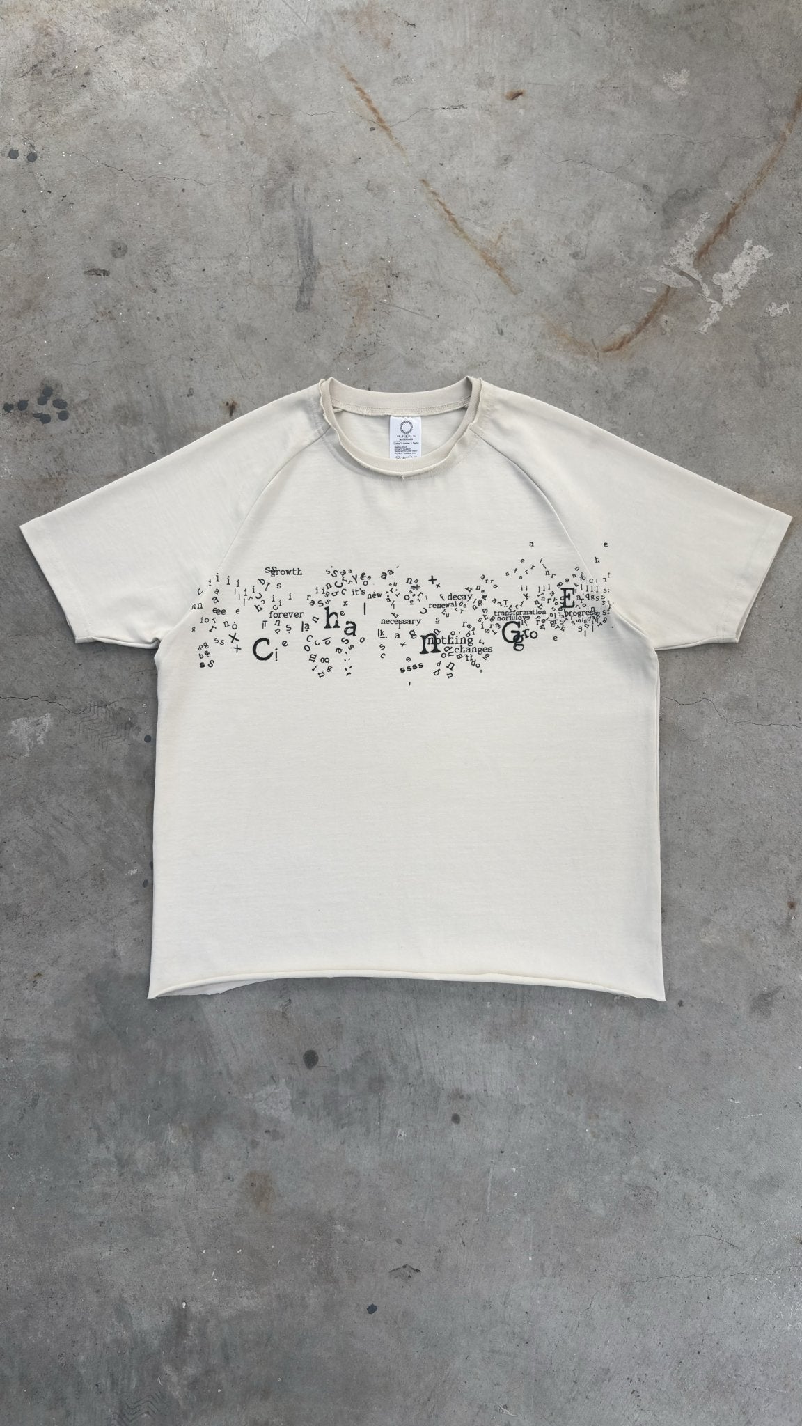 A zoomed-out overhead flat lay shot capturing the complete silhouette of the Poem Tee spread wide on a raw concrete garage floor. The full-profile view highlights the 325 GSM French Terry fleece-cotton construction with its saddled-raglan slim drop-shoulder sleeve paneling and custom garment proportions. Key details visible across the piece include the prominent direct-to-garment torso print, stacked raw-edge ribbed neckline, and intricate hand-cut Dremel micro-distressing along the hems and collar line.