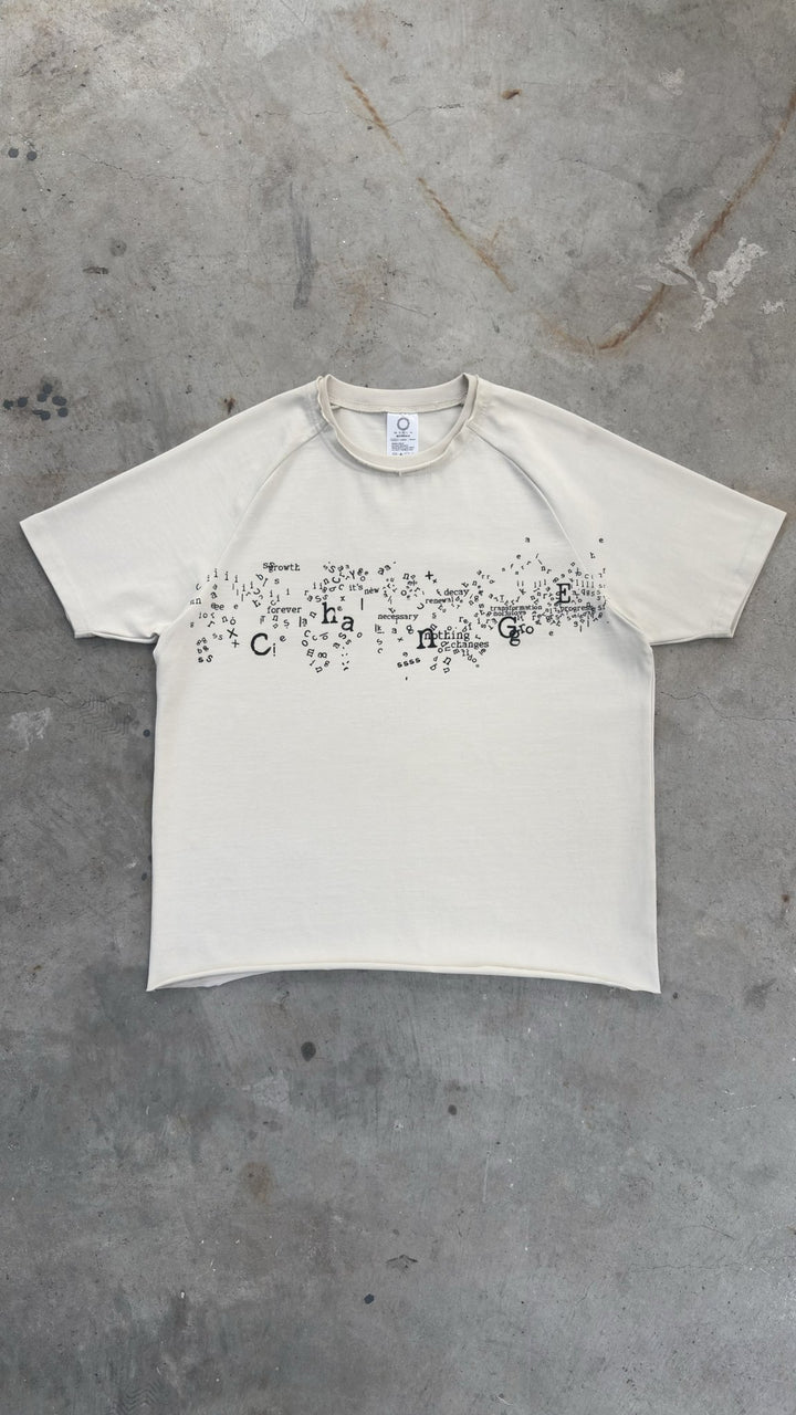 A zoomed-out overhead flat lay shot capturing the complete silhouette of the Poem Tee spread wide on a raw concrete garage floor. The full-profile view highlights the 325 GSM French Terry fleece-cotton construction with its saddled-raglan slim drop-shoulder sleeve paneling and custom garment proportions. Key details visible across the piece include the prominent direct-to-garment torso print, stacked raw-edge ribbed neckline, and intricate hand-cut Dremel micro-distressing along the hems and collar line.