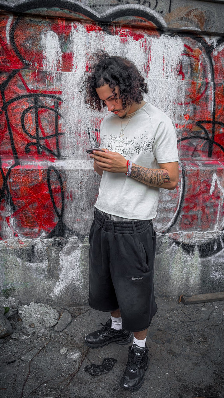 A full-body editorial portrait of the 6'1" light-skinned male model with curly hair standing outdoors. He wears the Poem Tee (325 GSM French Terry fleece-cotton with direct-to-garment torso print and stacked raw-edge ribbed neckline) paired with dark unreleased Y2K 3/4 length shorts and Balenciaga Runner sneakers. The wide shot captures the complete oversized drape, saddled-raglan drop-shoulder sleeve paneling, and overall footwear styling.