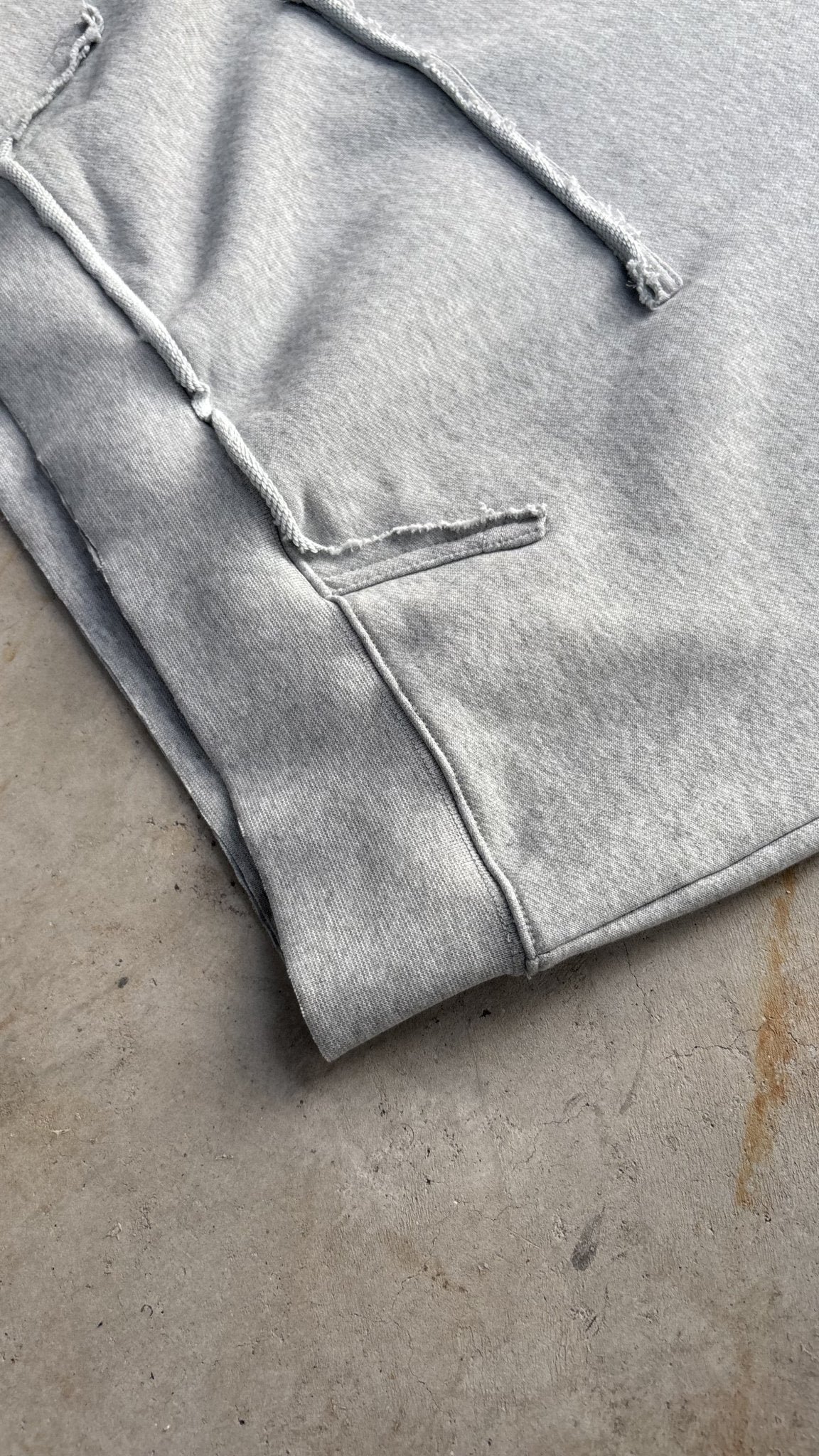 A macro top-down flat lay close-up focused on the deconstructed "ghost" kangaroo pocket of the Emo Hoodie, lying on a raw concrete garage floor. The shot emphasizes the frayed, hand-distressed perimeter of the torn pocket border, highlighting exposed French Terry interior loops, frayed raw thread edges, and the tonal silk-threaded torso embroidery adjacent to the pocket seam.
