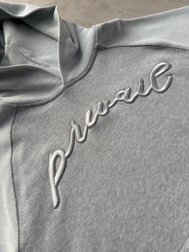 A macro top-down flat lay close-up focused on the center chest of the Emo Hoodie, resting on a raw concrete garage floor. The shot highlights the fine grey silk-threaded tonal embroidery precisely stitched across the heathered grey cotton torso, detailing the sheen and density of the thread work contrasting against the soft texture of the heavyweight cotton fleece fabric.