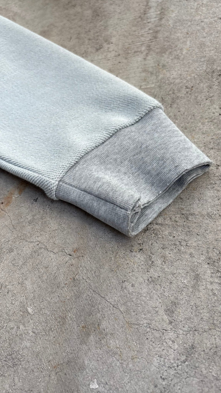 A macro flat lay close-up capturing the wrist opening of the Emo Hoodie on a raw concrete garage floor, detailing the material transition where the French Terry loop sleeve fabric joins the heathered grey cotton blend wrist cuff. The close-up showcases the 2x2 rib-knit cuff finishing, raw open-hem structural detailing, and the subtle deconstructed aesthetic surrounding the wrist construction.