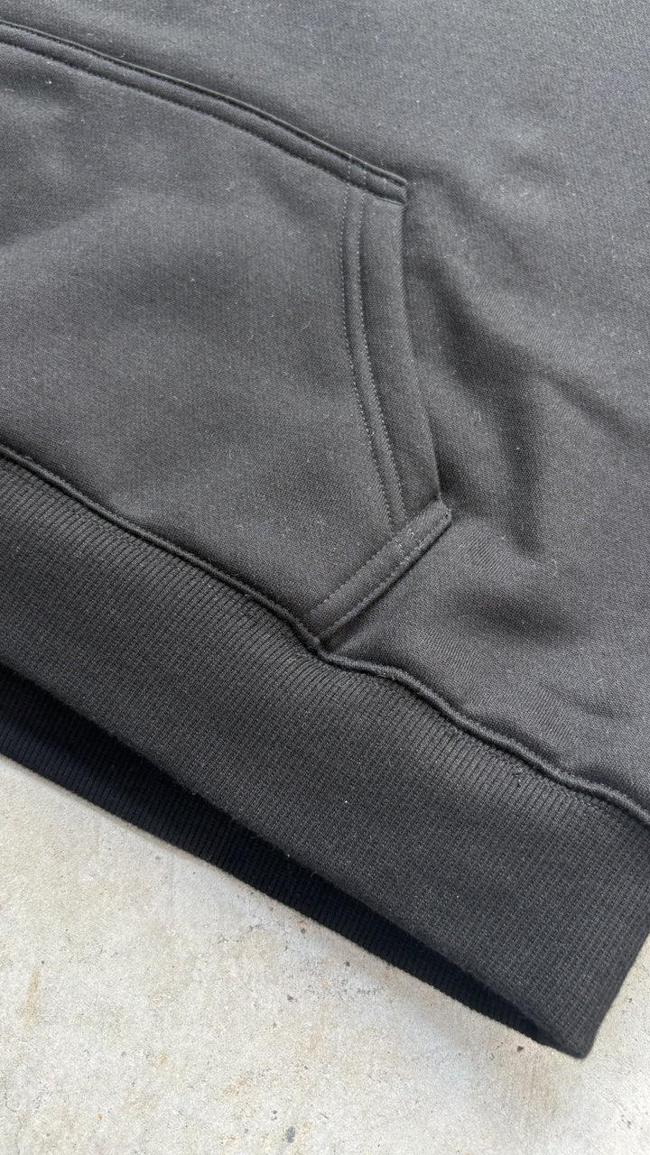 A macro flat lay close-up shot focused on the kangaroo pocket and bottom hem of the black Onyx Hoodie, resting on a concrete garage floor with the bottom edge facing the bottom-left corner. The tight frame highlights the intricate seam stitching, durable reinforced pocket edges, and 1x1 rib-knit waistband construction, detailing the premium finish of the 420 GSM heavyweight cotton-blend fabric alongside silk-threaded tonal embroidery accents.