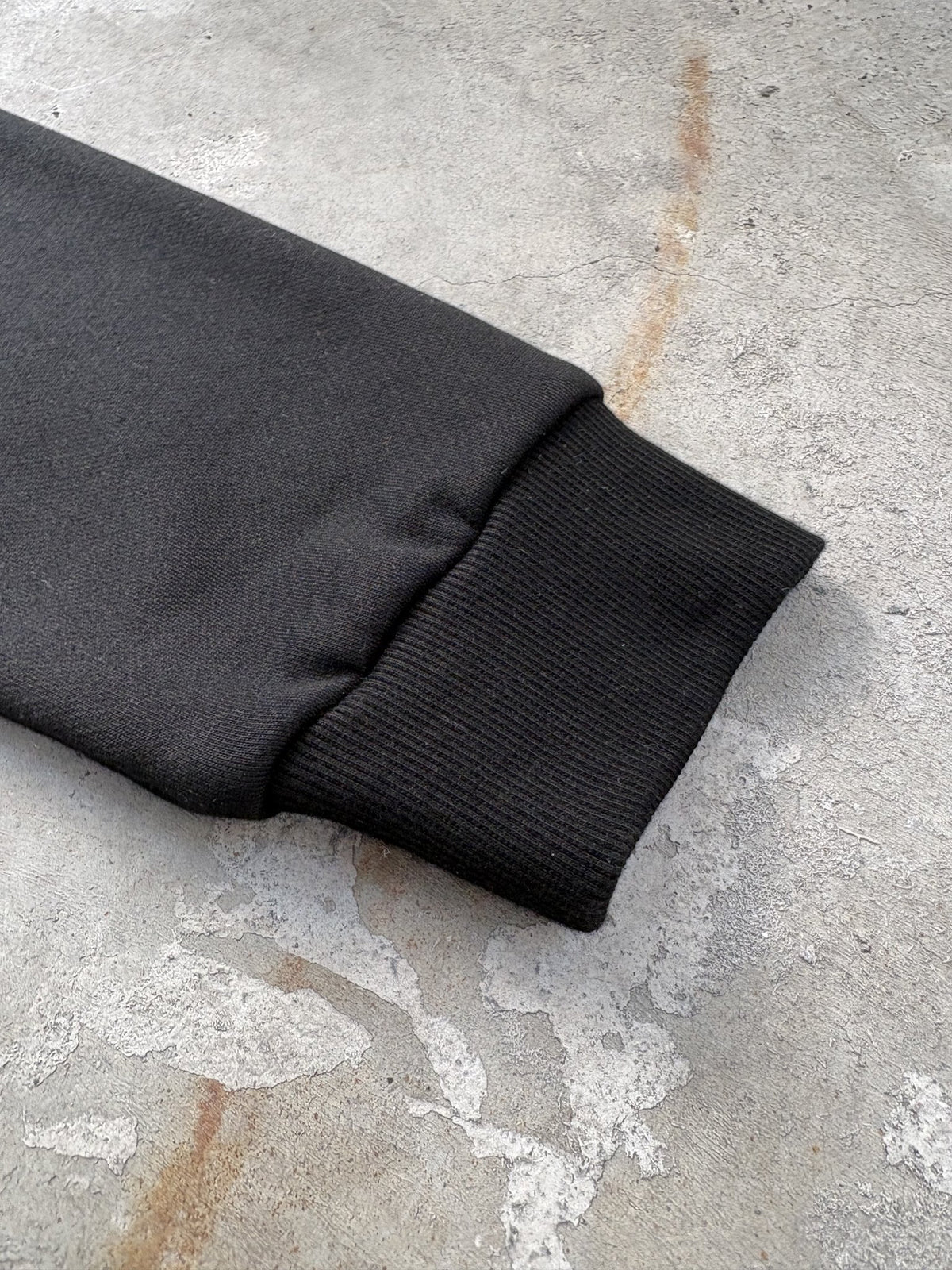 A macro flat lay close-up shot focused on a single wrist cuff of the black Onyx Hoodie, resting on a concrete garage floor. The tight frame highlights the 1x1 rib-knit cuff structure, precision seam stitching, and the subtle textural contrast between the elastic ribbing and the 420 GSM heavyweight cotton-blend fleece body, showcasing the meticulous garment construction and silk-threaded tonal embroidery details near the wrist.