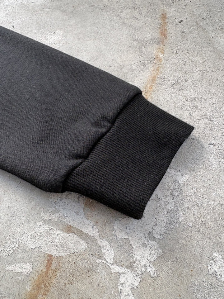 A macro flat lay close-up shot focused on a single wrist cuff of the black Onyx Hoodie, resting on a concrete garage floor. The tight frame highlights the 1x1 rib-knit cuff structure, precision seam stitching, and the subtle textural contrast between the elastic ribbing and the 420 GSM heavyweight cotton-blend fleece body, showcasing the meticulous garment construction and silk-threaded tonal embroidery details near the wrist.