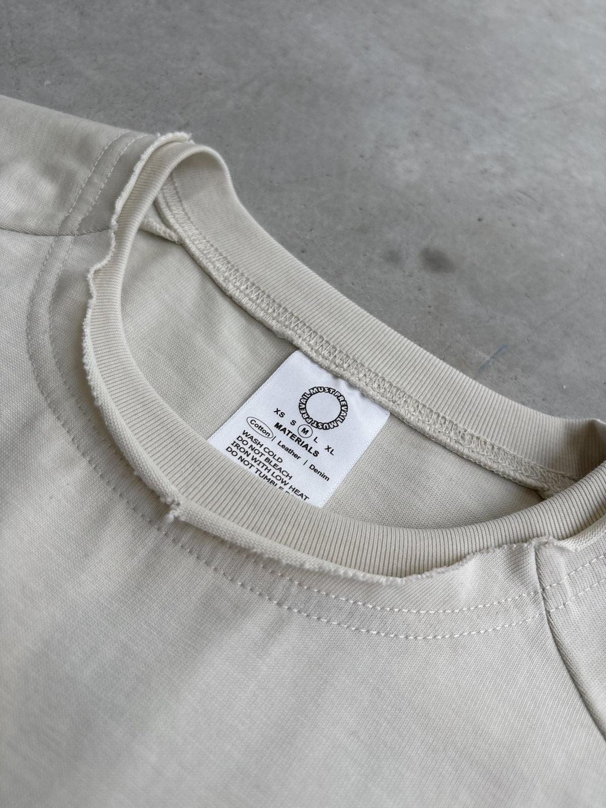 A macro top-down flat lay shot focused on the neckline and collar structure of the Poem Tee, laid flat on a raw concrete garage floor. The close-up view highlights the stacked raw-edge ribbed double-collar neckline, hand-cut Dremel micro-distressing details, and the custom cream-colored neck tag. The tight framing captures the 325 GSM French Terry fleece-cotton fabric texture, intricate seam stitching, and the saddled-raglan slim drop-shoulder sleeve paneling where the shoulder connects to the torso print.