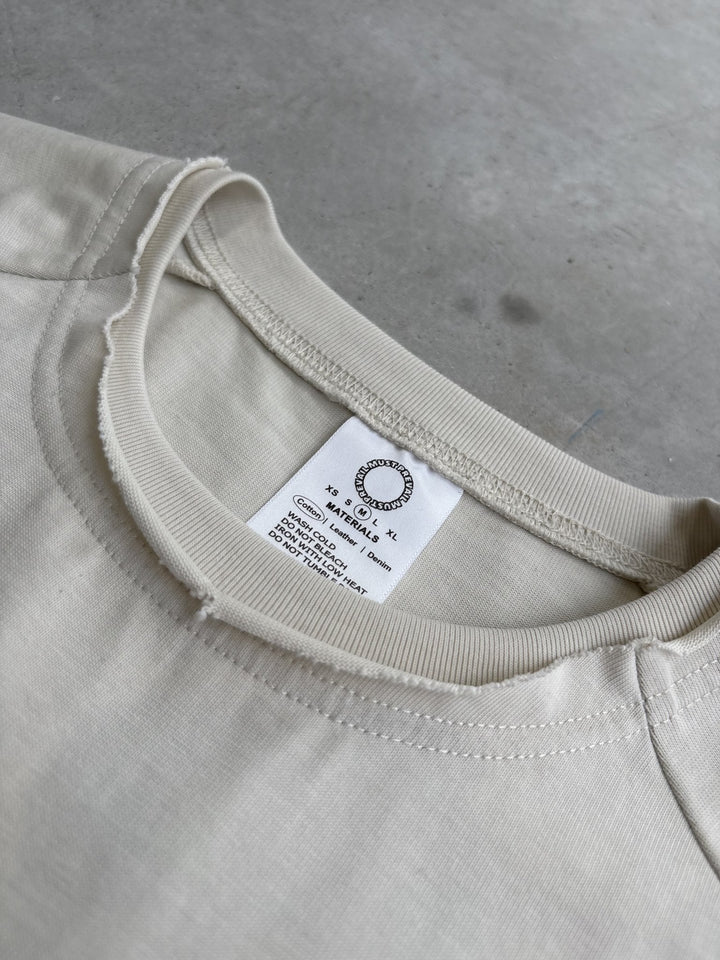 A macro top-down flat lay shot focused on the neckline and collar structure of the Poem Tee, laid flat on a raw concrete garage floor. The close-up view highlights the stacked raw-edge ribbed double-collar neckline, hand-cut Dremel micro-distressing details, and the custom cream-colored neck tag. The tight framing captures the 325 GSM French Terry fleece-cotton fabric texture, intricate seam stitching, and the saddled-raglan slim drop-shoulder sleeve paneling where the shoulder connects to the torso print.