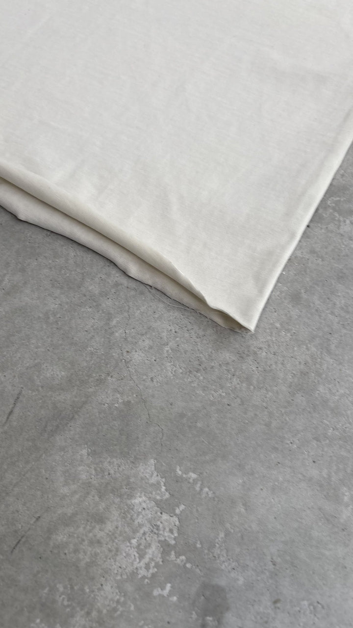 A macro top-down flat lay close-up of the Poem Tee's bottom hem on a raw concrete garage floor, positioned diagonally across the frame. This shot is the reverse angle of the first bottom hem close-up, showcasing the meticulous construction of the 325 GSM French Terry fleece-cotton fabric, the evenness of the double-needle hem stitching, and the distinct, raw texture of the Dremel micro-distressing from the opposite side.