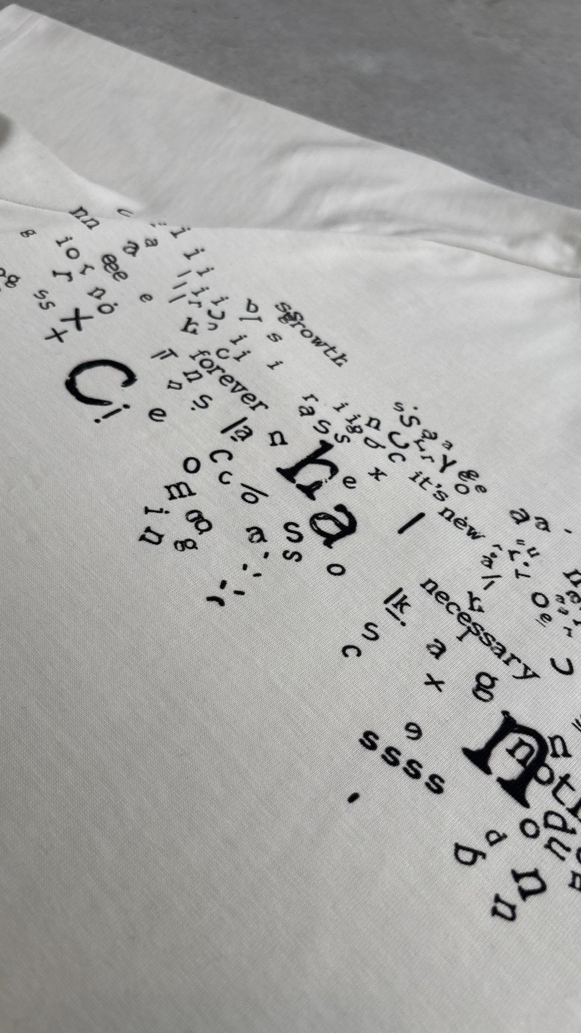 A macro top-down flat lay close-up of the Poem Tee's torso print, specifically focusing on the area near the left shoulder seam where the print meets the distinct saddled-raglan panel. This image showcases the precise alignment of the direct-to-garment graphics with the complex garment construction and the transition between the printed and unprinted 325 GSM French Terry fleece-cotton fabric on the raw concrete garage floor.