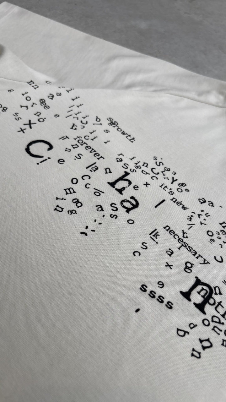 A macro top-down flat lay close-up of the Poem Tee's torso print, specifically focusing on the area near the left shoulder seam where the print meets the distinct saddled-raglan panel. This image showcases the precise alignment of the direct-to-garment graphics with the complex garment construction and the transition between the printed and unprinted 325 GSM French Terry fleece-cotton fabric on the raw concrete garage floor.