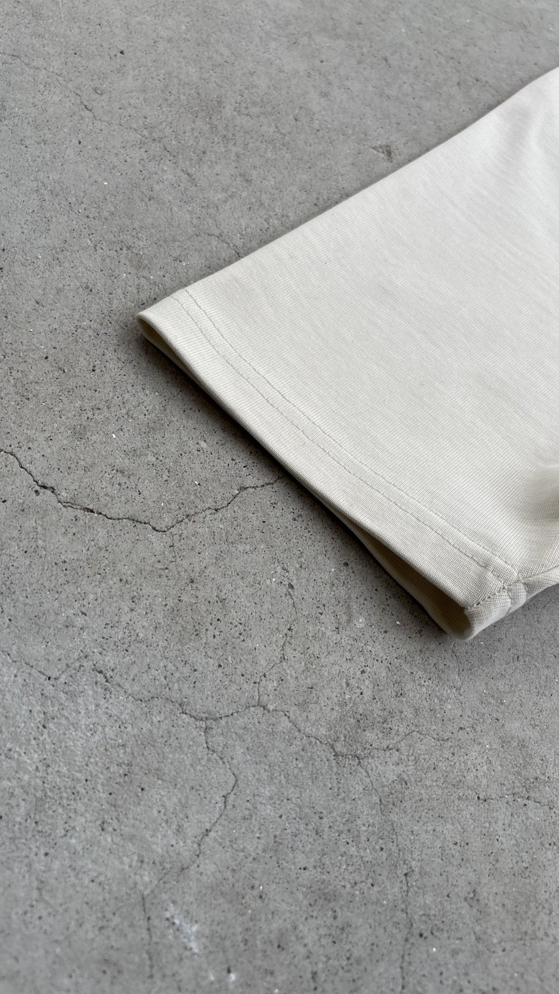 A top-down macro flat lay image focusing exclusively on the opening of the Poem Tee's sleeve, resting on a raw concrete garage floor. The shot avoids showing the main sleeve paneling, concentrating on the raw-edge hem finish, the precise single-needle stitching, and the distinct texture of the 325 GSM French Terry fleece-cotton, with subtle Dremel micro-distressing along the hem.