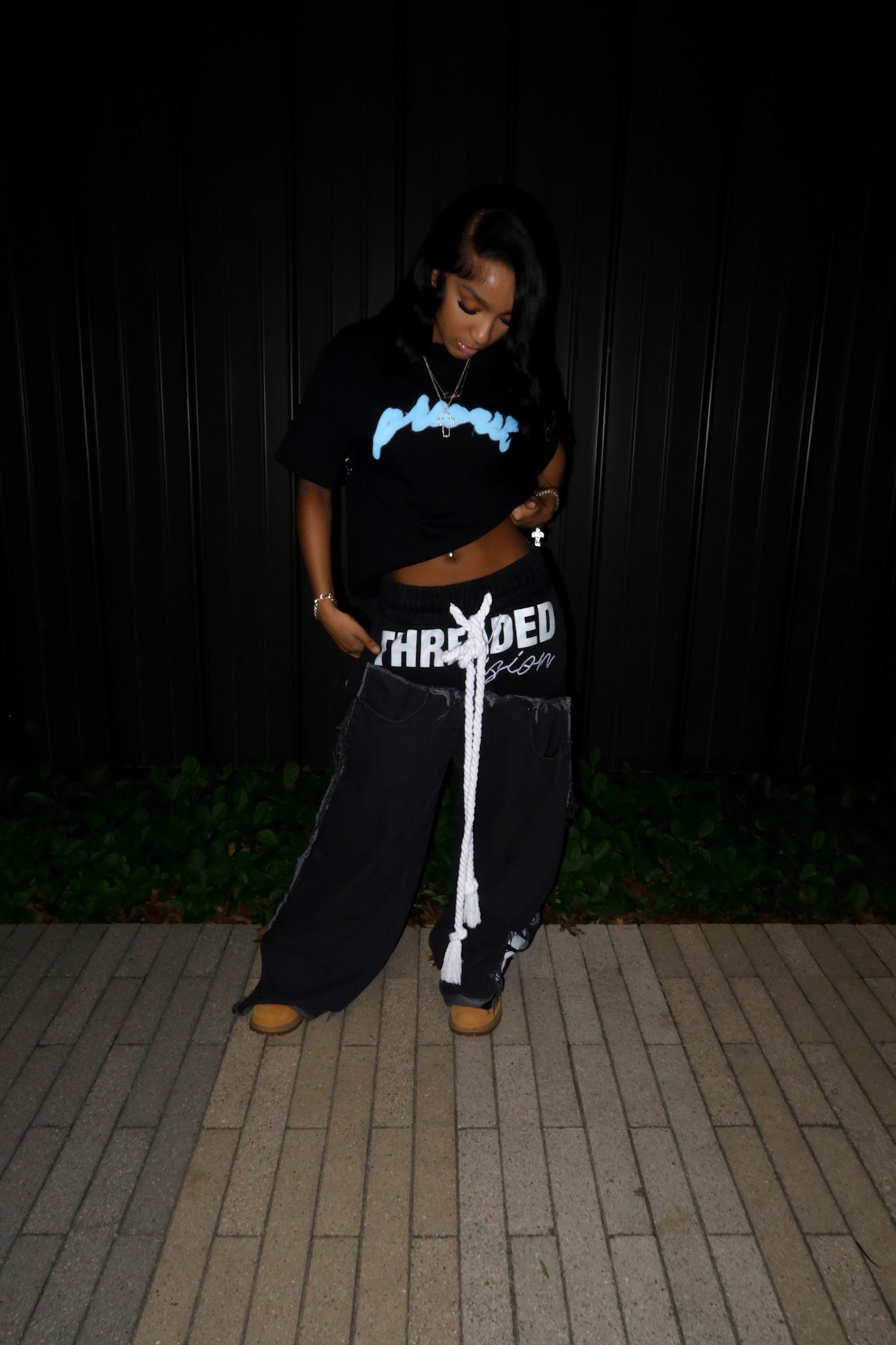 A dynamic lifestyle angle of a model in the MUST PREVAIL black statement tee. The tee is styled with a cropped hem to highlight the high-waisted wide-leg trousers and classic wheat Timberland boots. This flash-lit night shot showcases the oversized sleeves, the teal 'Prevail' DTG graphic, and the heavy architectural drape of the 350 GSM cotton.