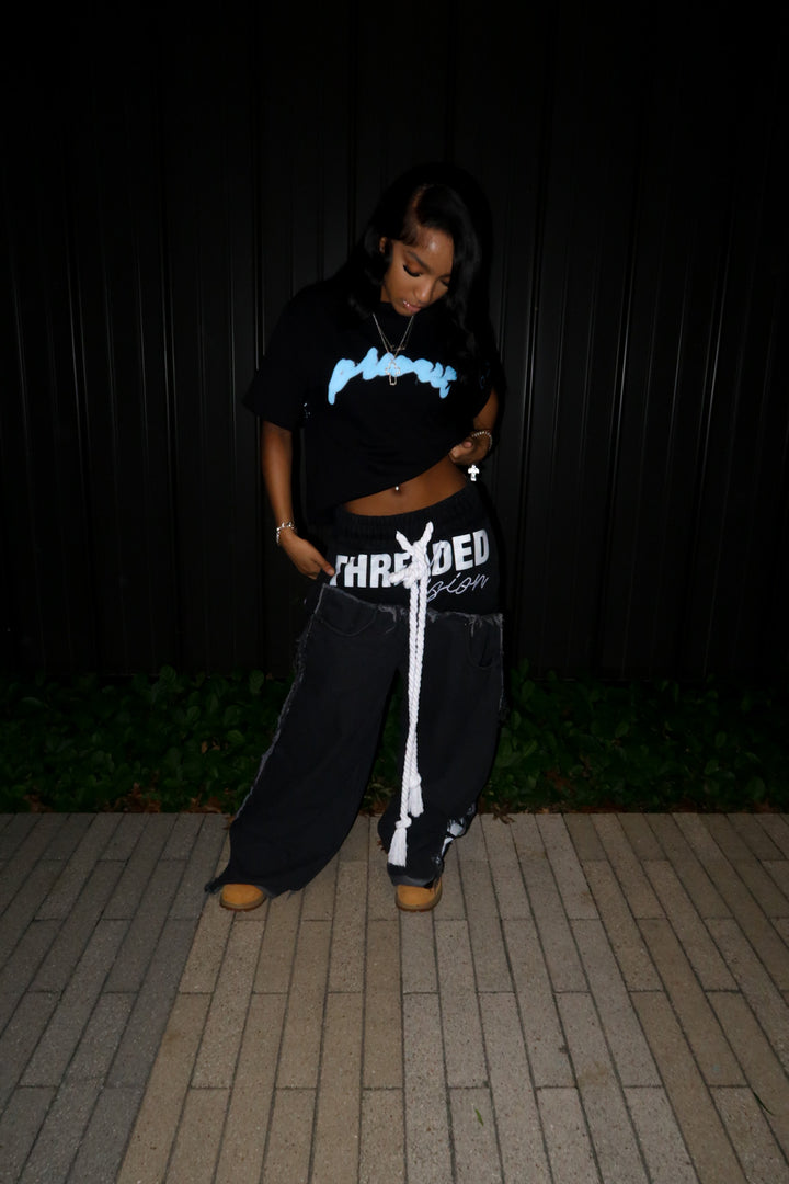 A dynamic lifestyle angle of a model in the MUST PREVAIL black statement tee. The tee is styled with a cropped hem to highlight the high-waisted wide-leg trousers and classic wheat Timberland boots. This flash-lit night shot showcases the oversized sleeves, the teal 'Prevail' DTG graphic, and the heavy architectural drape of the 350 GSM cotton.