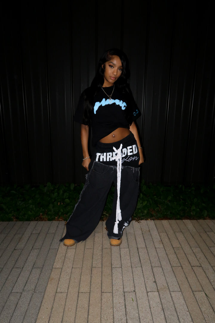 A female model wearing the MUST PREVAIL black statement tee styled with a cropped hem. The image features the teal blue 'Prevail' DTG graphic and is paired with oversized wide-leg black trousers and classic timberland “wheat” boots. This night-time flash photography highlights the boxy silhouette and urban drape of the heavyweight cotton.